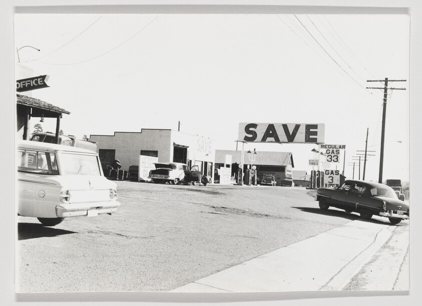 Large Save sign towers over a mid-century gas station with cars and gas price signs.