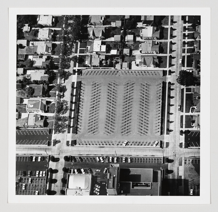 Aerial view showing a large empty parking lot with lined spaces surrounded by houses and streets.