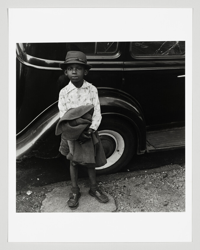 A black and white photograph of a young boy standing beside a vintage car, wearing an oversized hat and holding a coat. He appears to be looking directly at the camera with a serious expression.