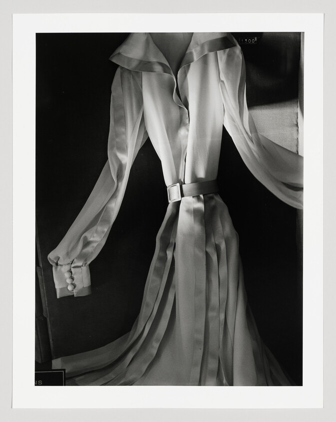 A black and white photograph of an elegant, flowing dress with long sleeves and a belted waist, displayed on a mannequin against a dark background. The dress features a ruffled collar and the fabric appears light and sheer, suggesting movement. The image is framed with a white border.