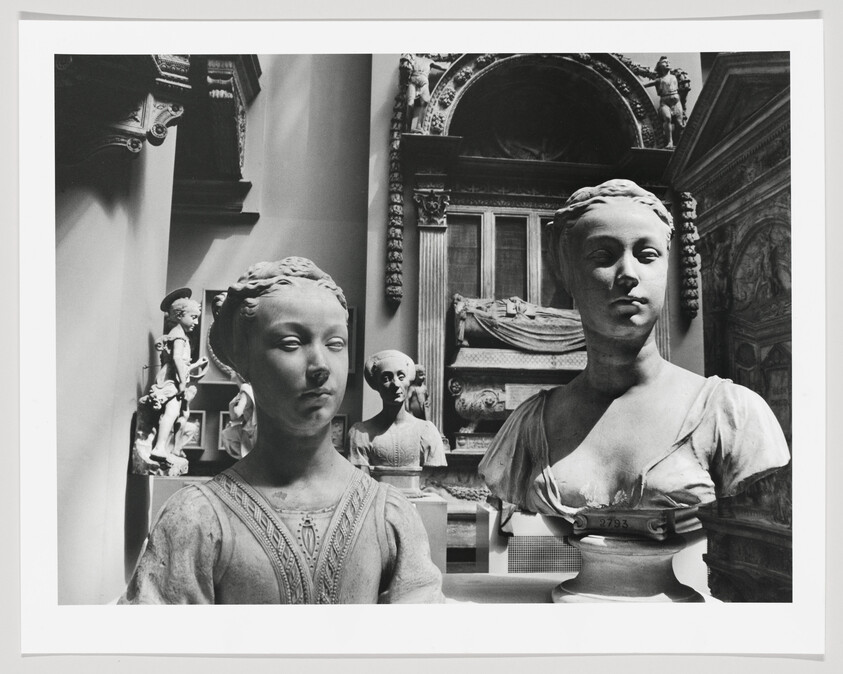Black and white photograph of two classical bust sculptures prominently displayed in a museum setting with architectural details and additional sculptures in the background.