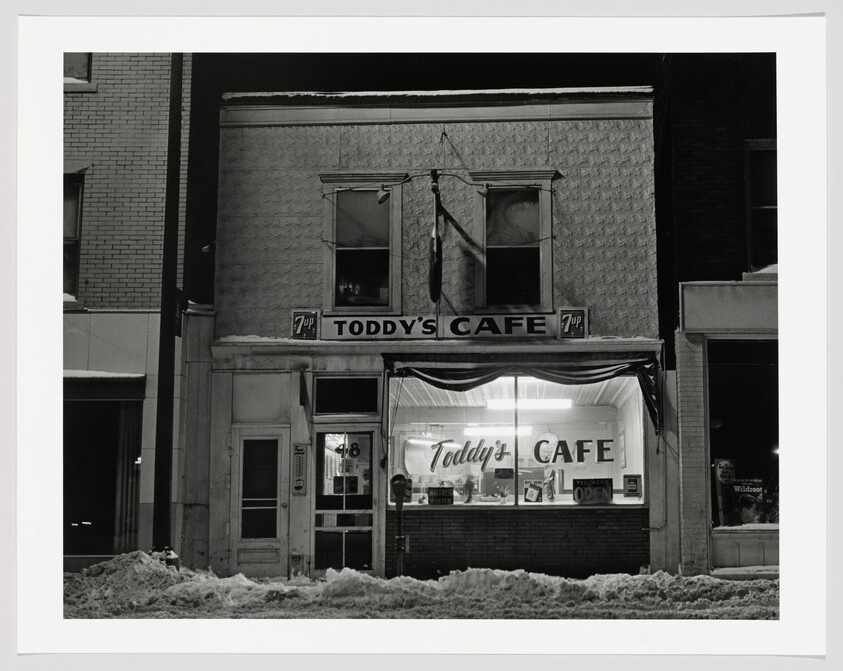 A black and white photograph of an old two-story building at night with a sign reading "Toddy's Café." The ground floor has a lit storefront with neon signs in the windows, and the upper floor has two windows with closed curtains. There is snow piled up on the sidewalk in front of the building.