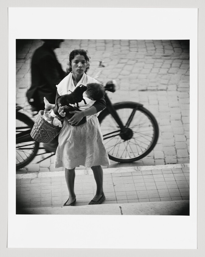 A black and white photograph capturing a young woman standing on a tiled pavement, holding a small black pig in one arm and a woven basket in the other. She is wearing a light-colored dress with a white shawl over her shoulders and looks directly at the camera with a neutral expression. In the blurred background, there is a partial view of a bicycle and the silhouette of a person walking by.