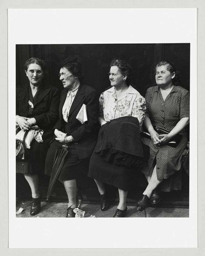 Four women sitting side by side against a dark background, with the second from the left having a noticeably smudged face. They are dressed in mid-20th-century attire, with three of them holding handbags and one holding a folded piece of paper. Their expressions range from neutral to slightly smiling, and one appears to be squinting in the sunlight.
