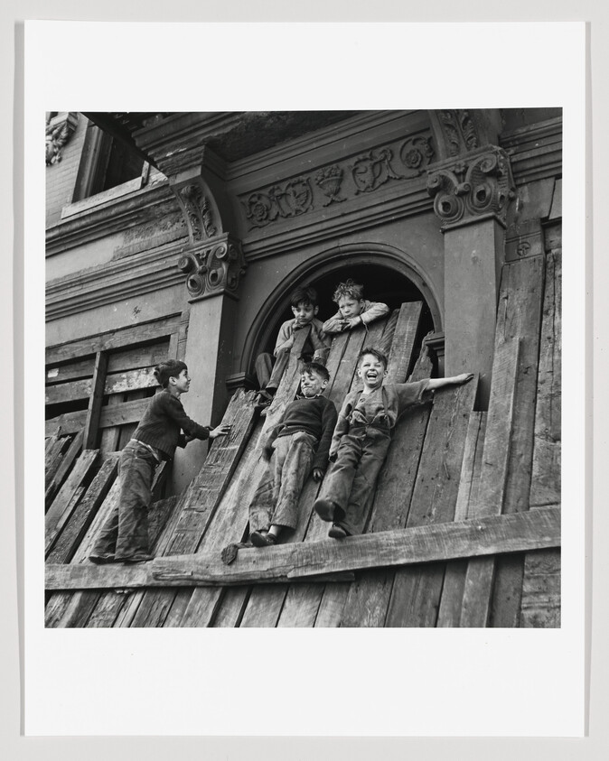 A black and white photograph showing a group of children playing on a makeshift slide made from planks of wood. The slide is positioned on a set of stairs leading to an ornate doorway of an old building. Two boys are sliding down the plank, one lying on his back and the other sitting, both with expressions of joy. Another boy is standing at the bottom of the slide, looking up, while two more children are watching from the doorway above. The children are dressed in casual, worn clothing, suggesting a bygone era.