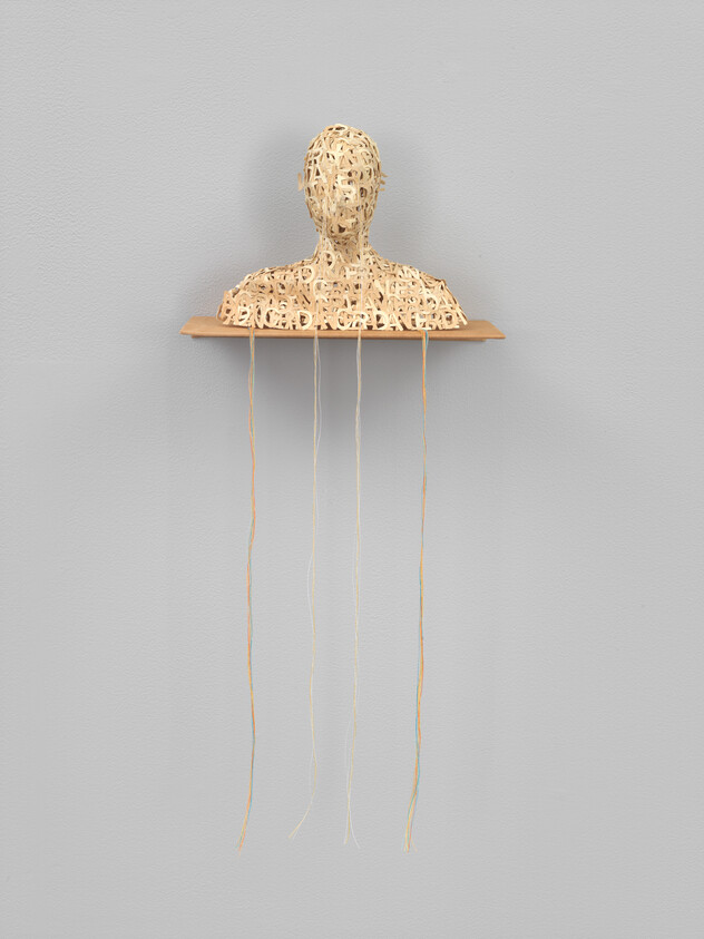 A woven human bust sits on a small shelf with long thin strings hanging below.