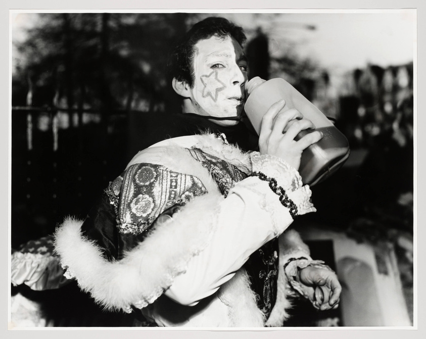 a white man in regalia grips a large bottle and stares at the camera