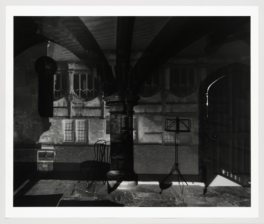 Stone column, empty chair, and music stand cast shadows under a vaulted ceiling near a partly open door.