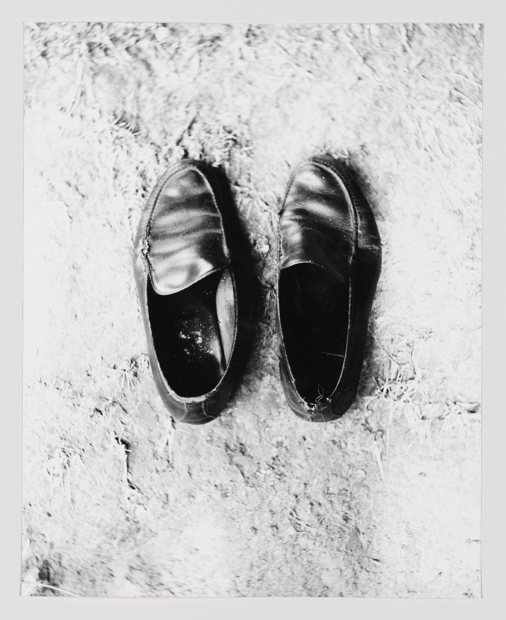 A pair of worn black leather slip-on shoes sitting side by side on rough ground.