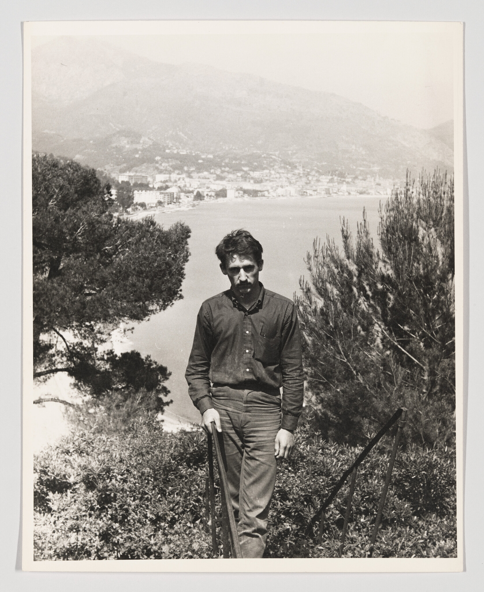 A man stands on a hillside path holding a railing, overlooking a coastal bay and town.