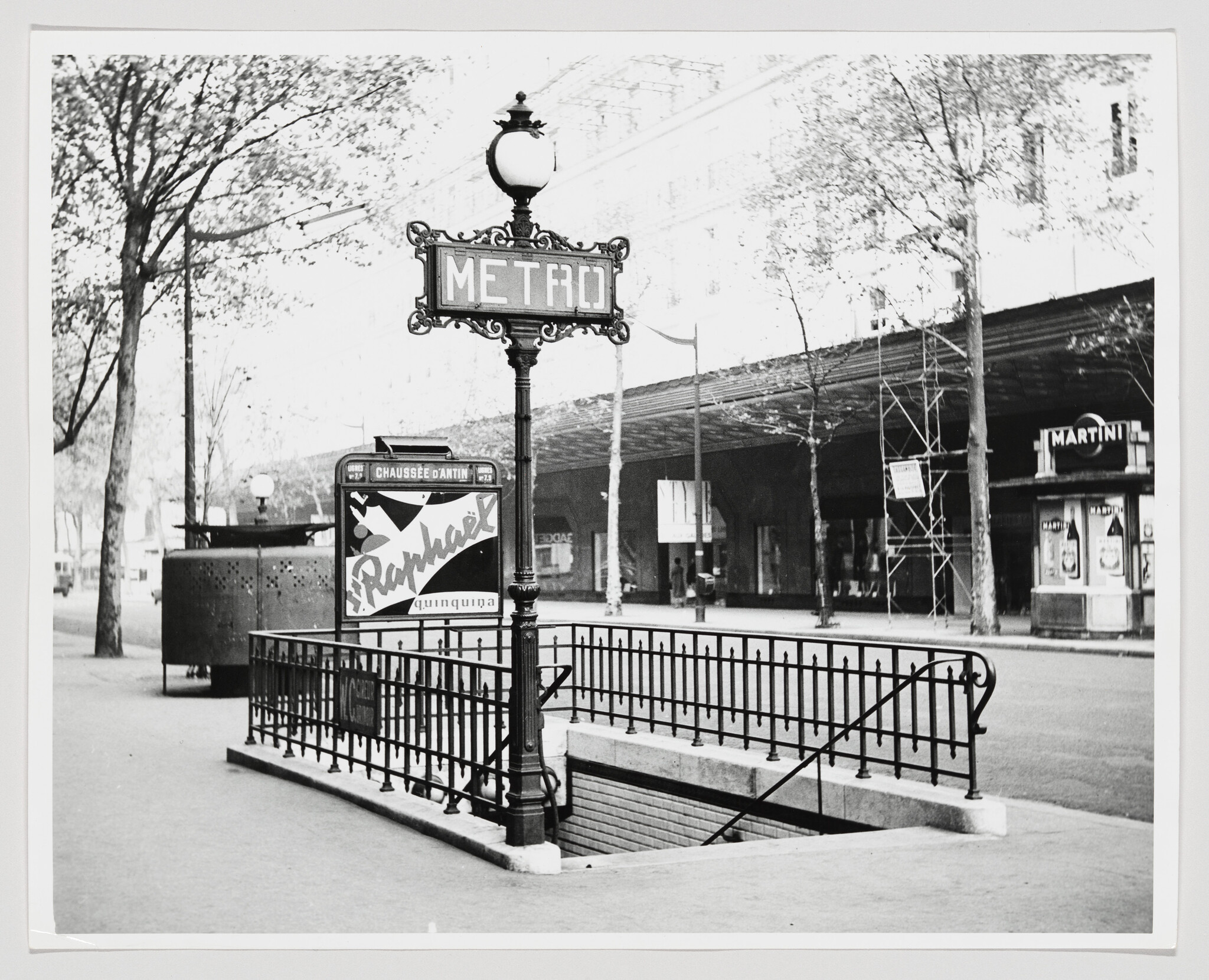 Ornate street lamp and METRO sign marking a fenced subway entrance on a tree-lined sidewalk.