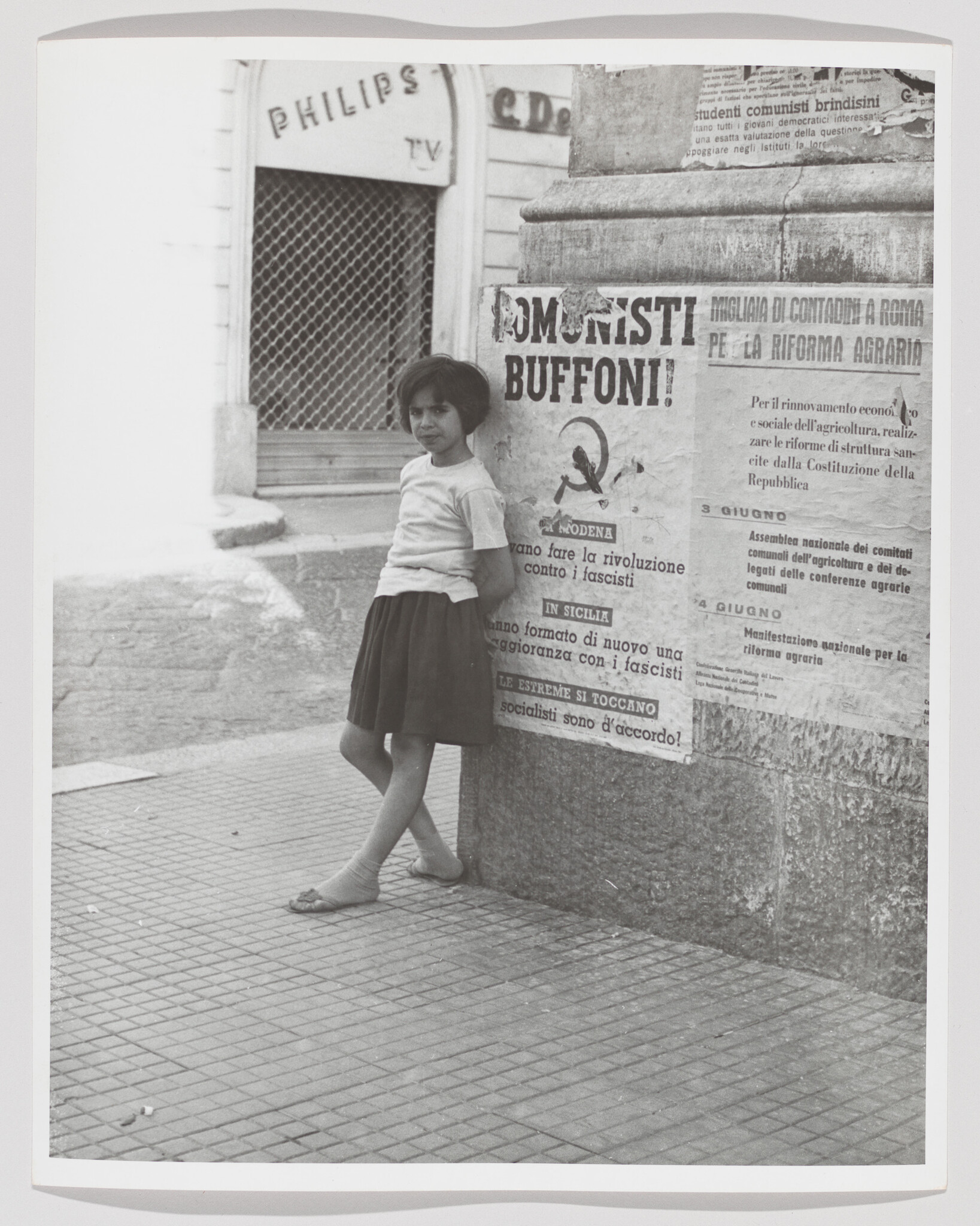 A young girl leans against a wall covered with political posters reading "Comunisti Buffoni".