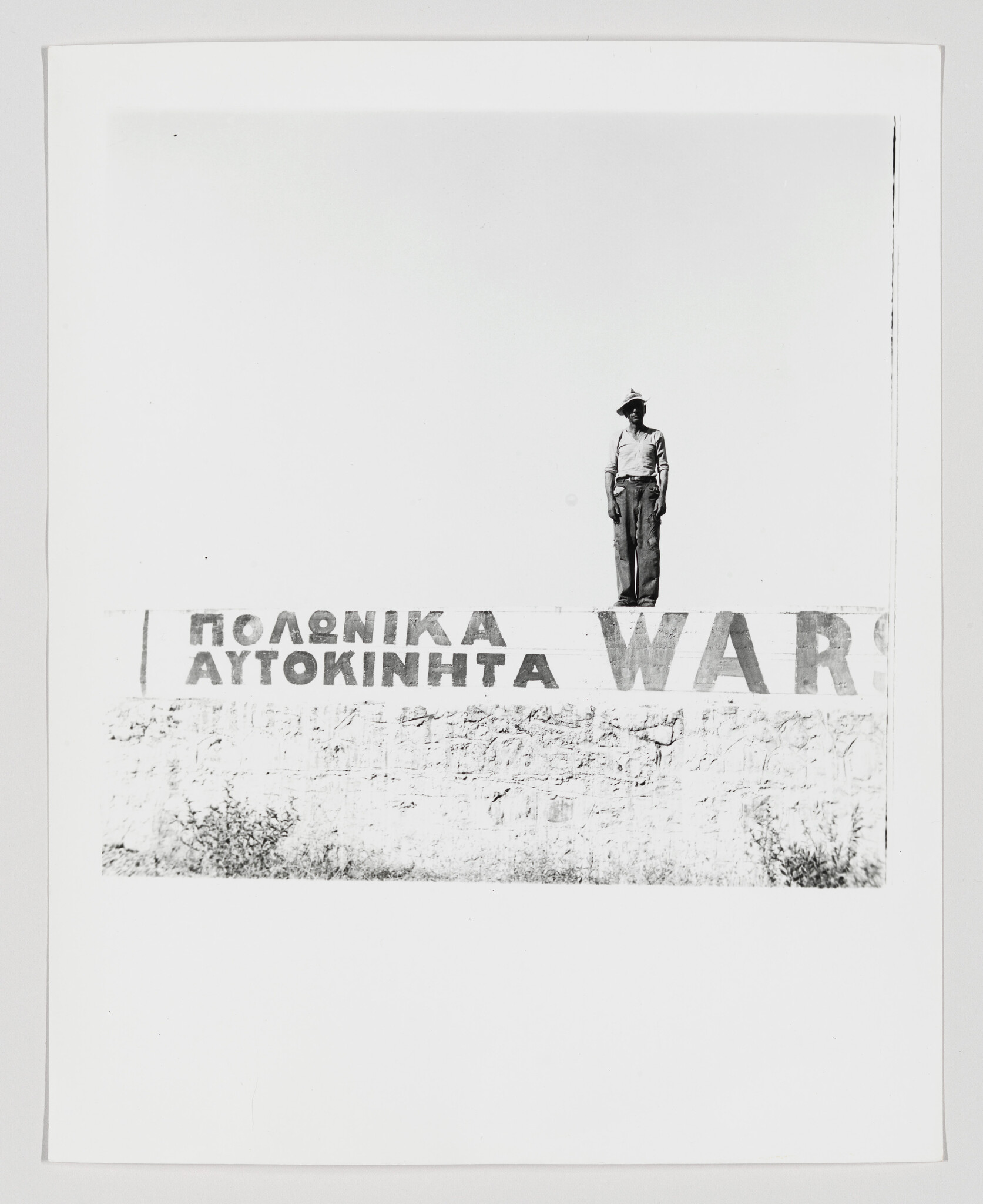 A man wearing a hat stands on a low wall above faded Greek advertising letters and WARS.