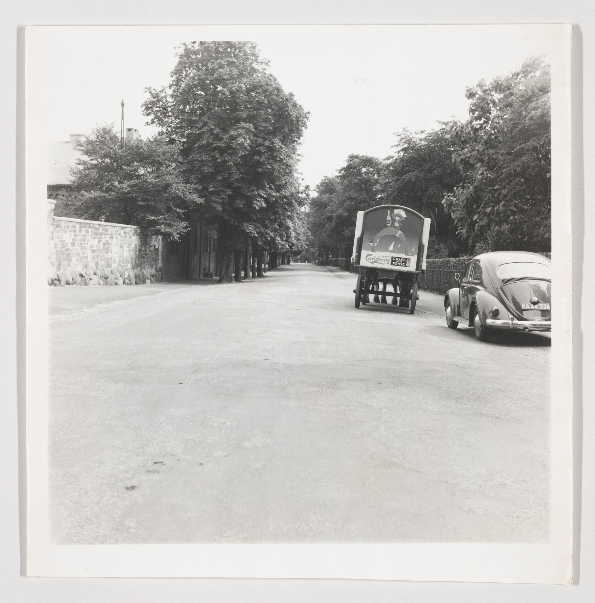 Horse-drawn beer delivery wagon and parked car stand on a quiet tree-lined street.