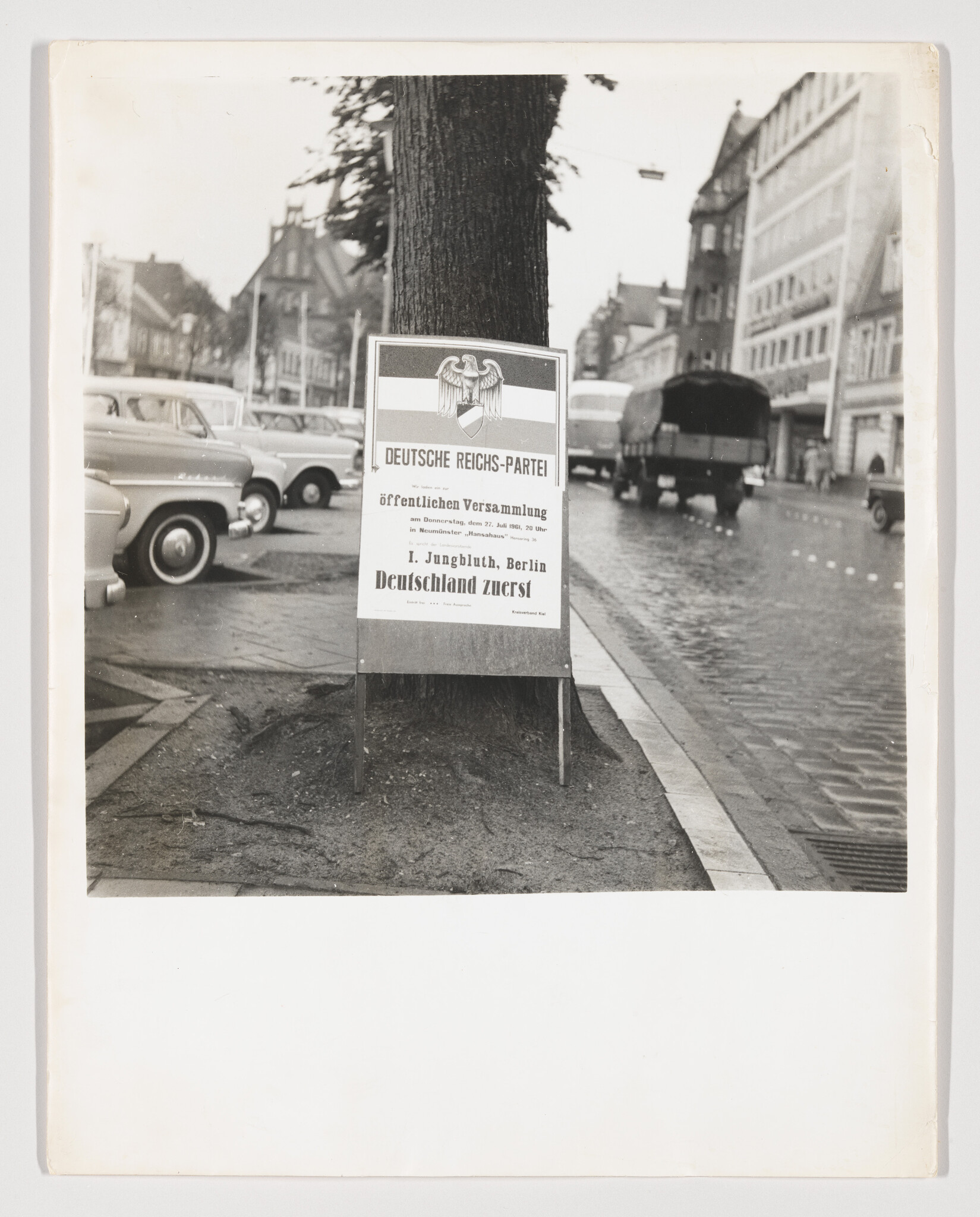 A political poster for the Deutsche Reichs-Partei is mounted on a tree beside a wet city street.