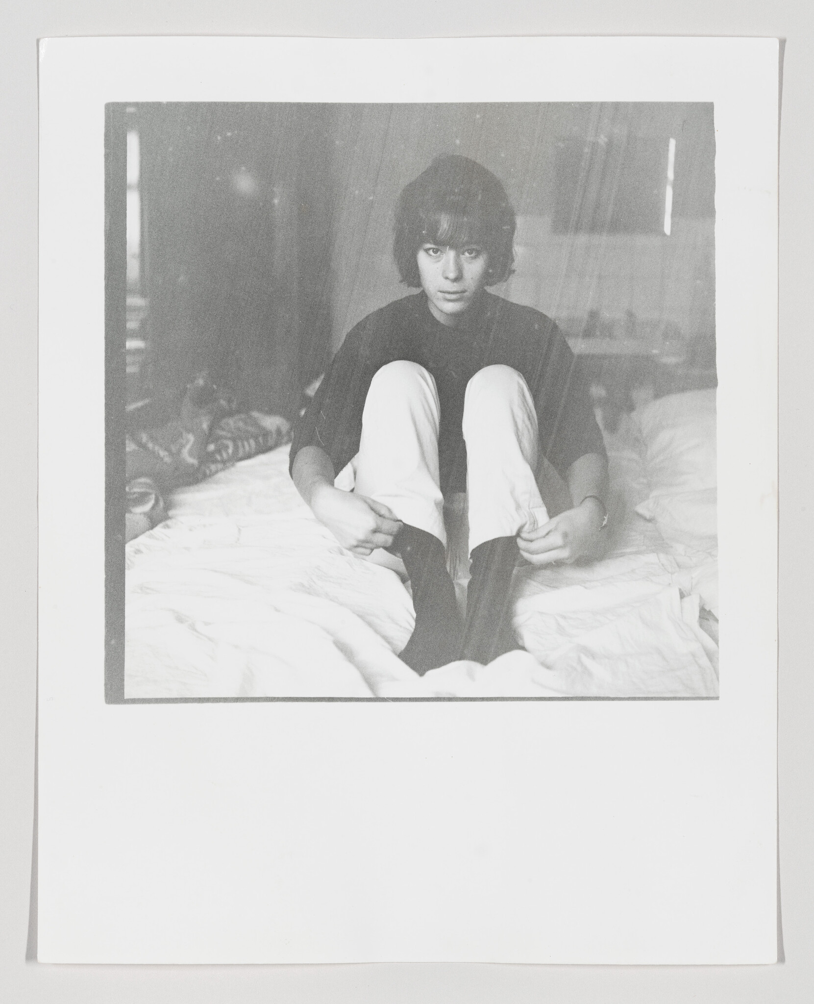 Teenager sits on a bed with knees up, pulling on dark socks while looking forward.