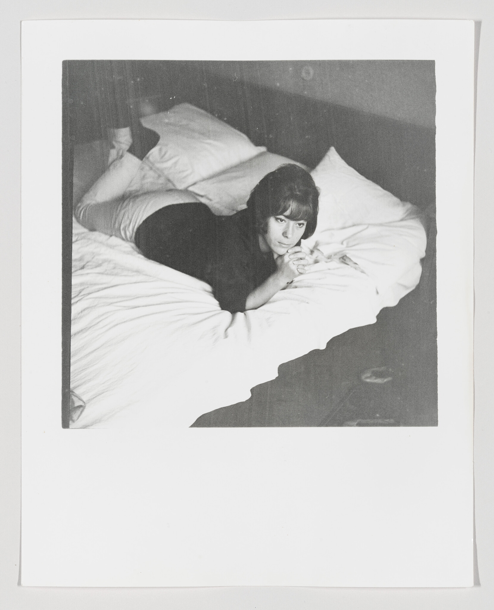 Woman lying on bed, propped on her elbows and looking thoughtfully toward camera.