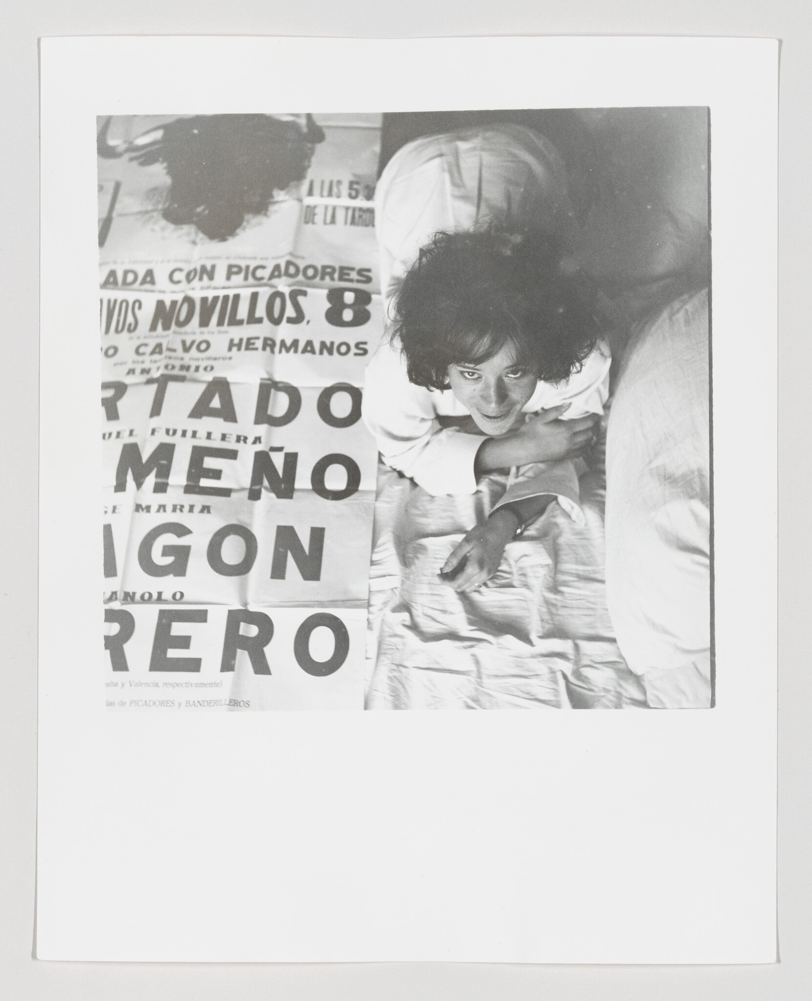 Young woman lies on a bed looking up beside a large printed poster.