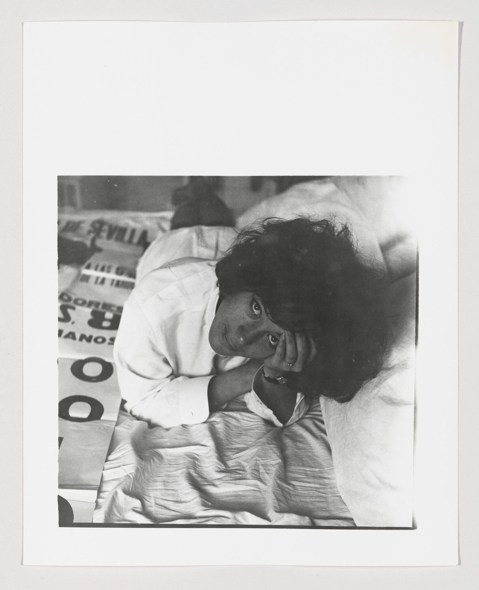 A person with dark curly hair lies on a bed, resting their head on one hand and looking up.