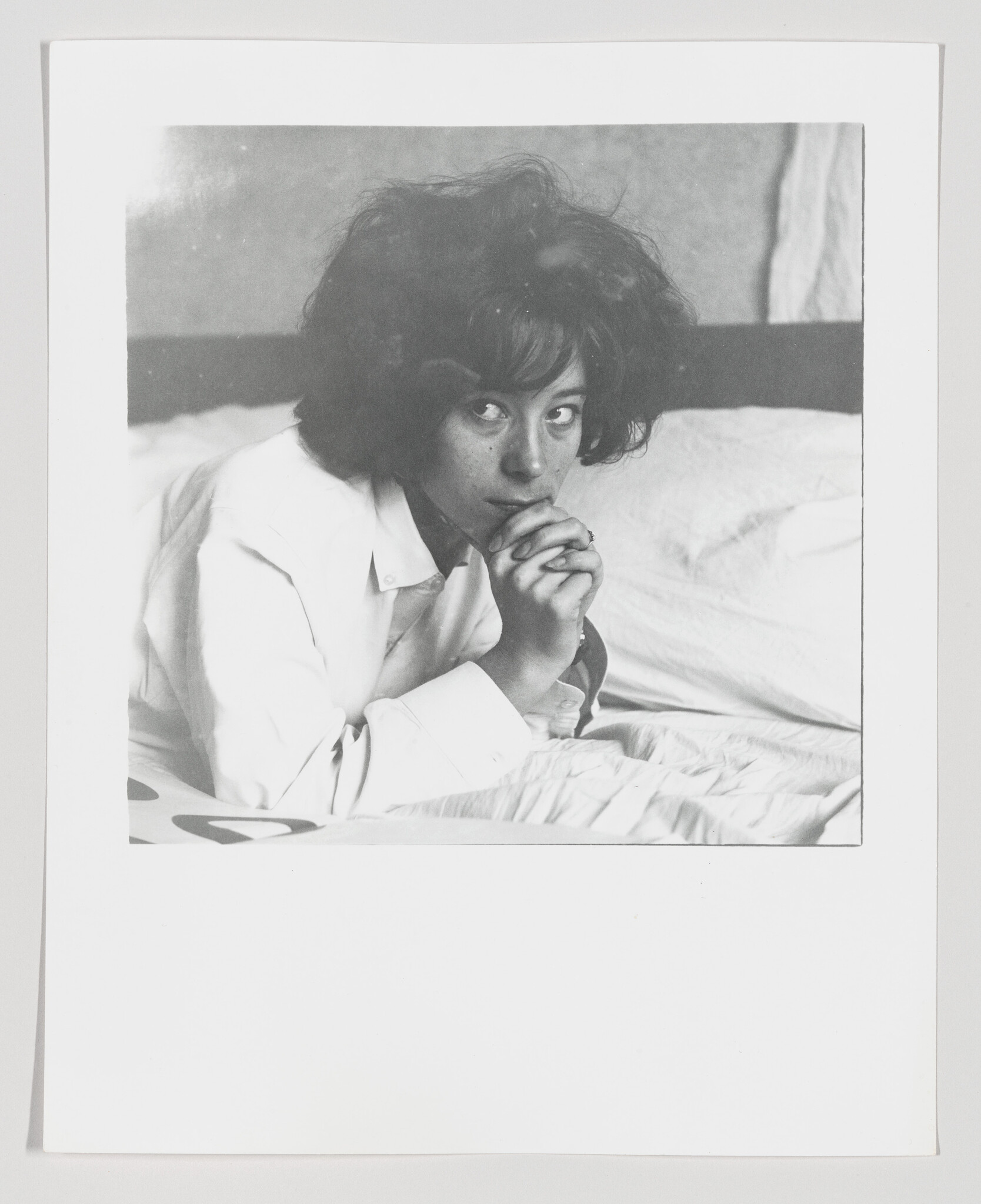 A young woman lies on a bed with her hands clasped under her chin, looking sideways.