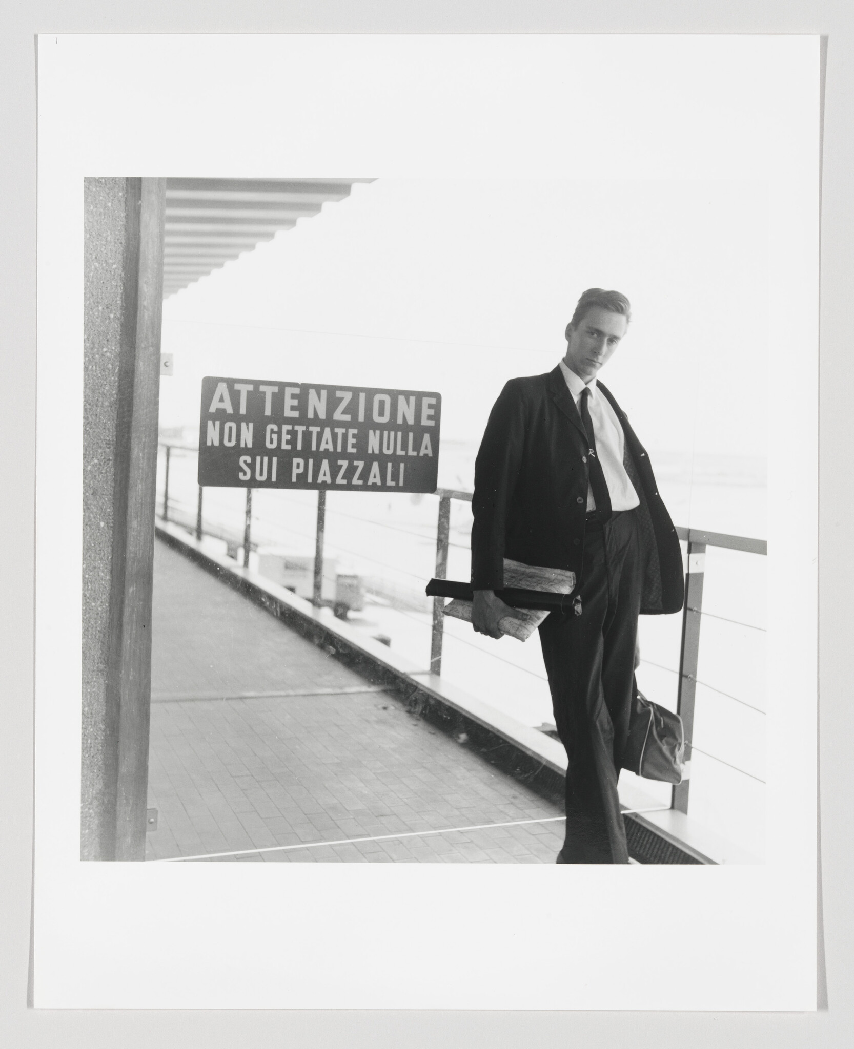 A young man in a suit leans on a railing holding papers beside a warning sign.