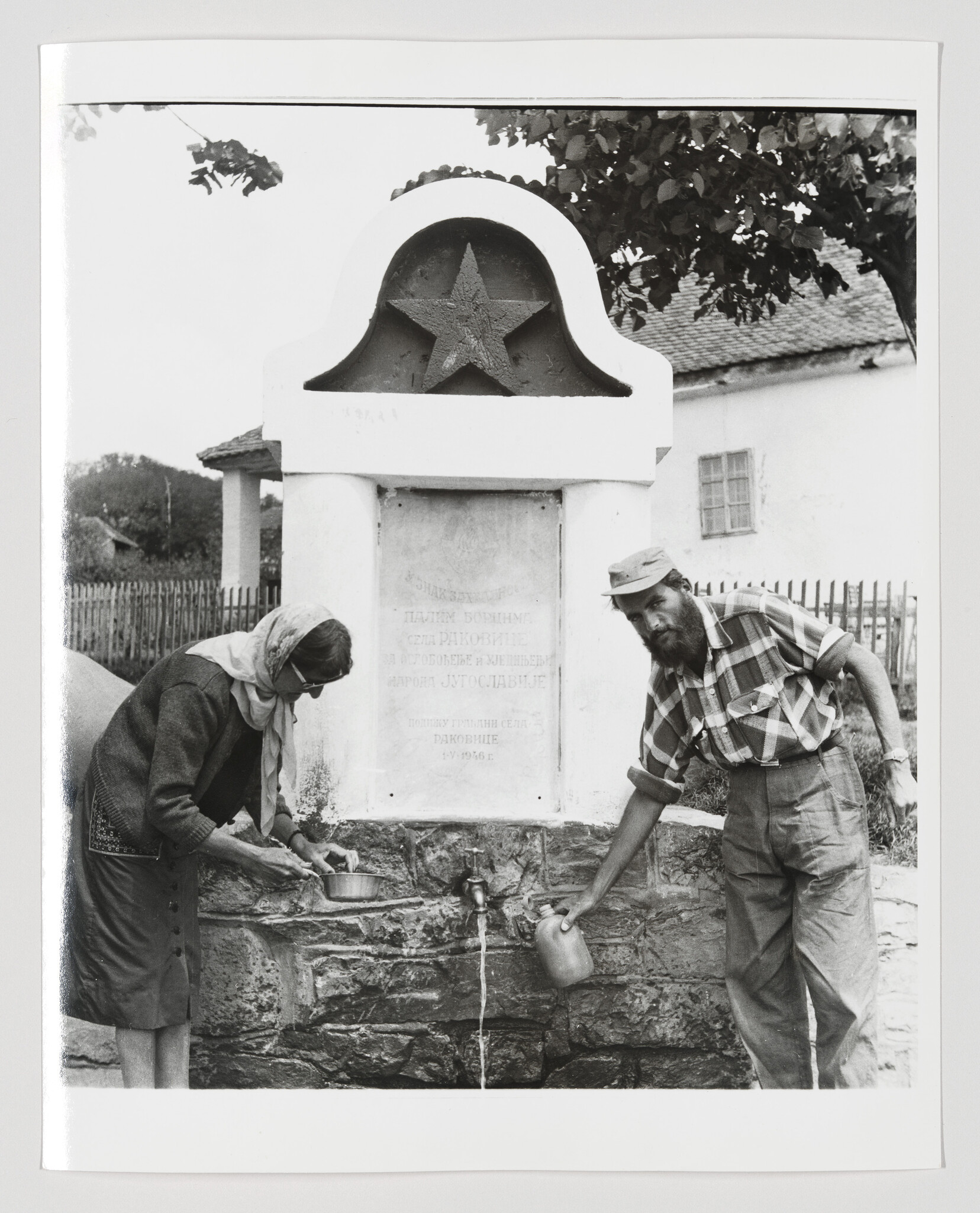 An elderly woman and a bearded man fill containers at a village water fountain with a star emblem.