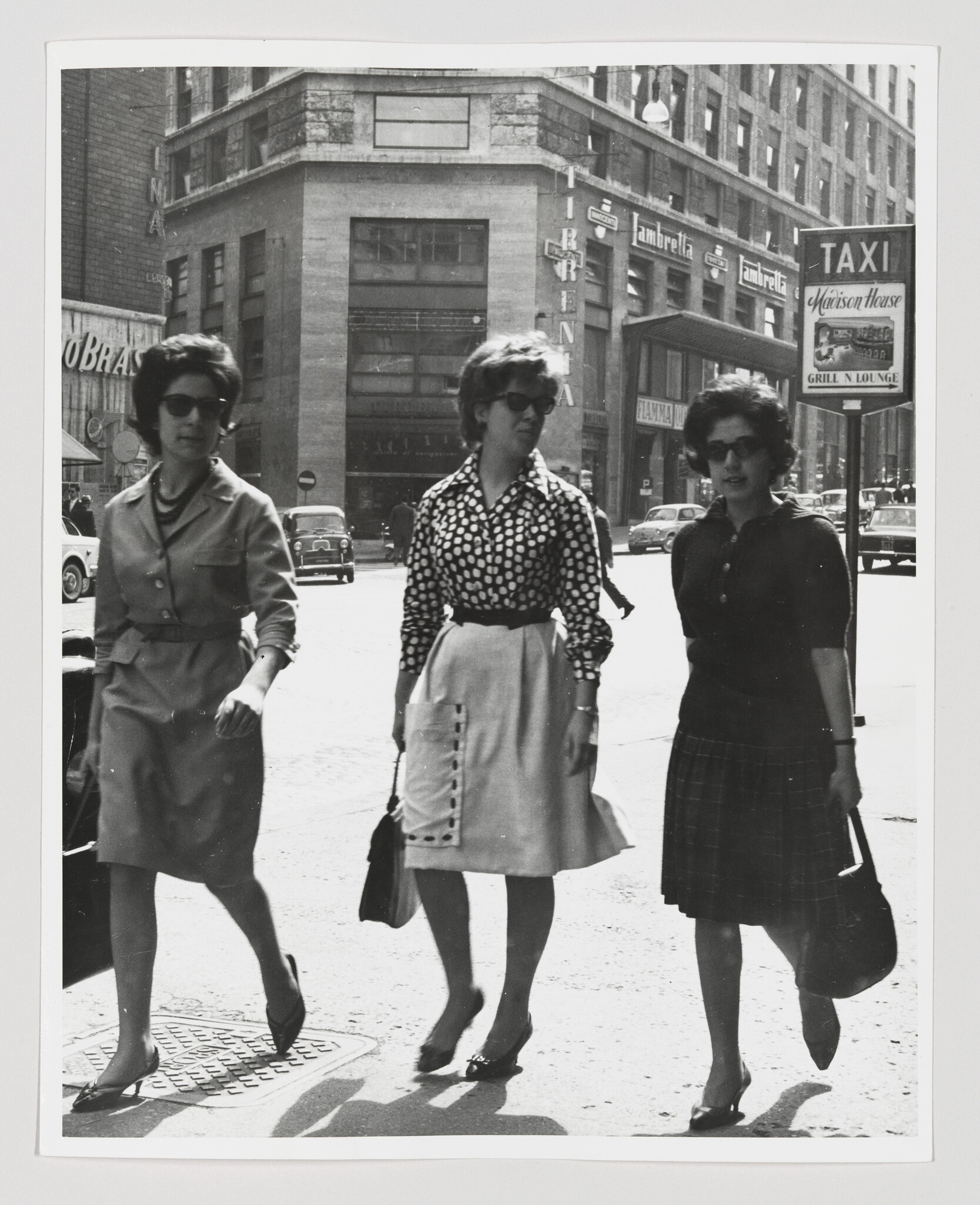 Three women in dresses and sunglasses walk together across a busy city street.