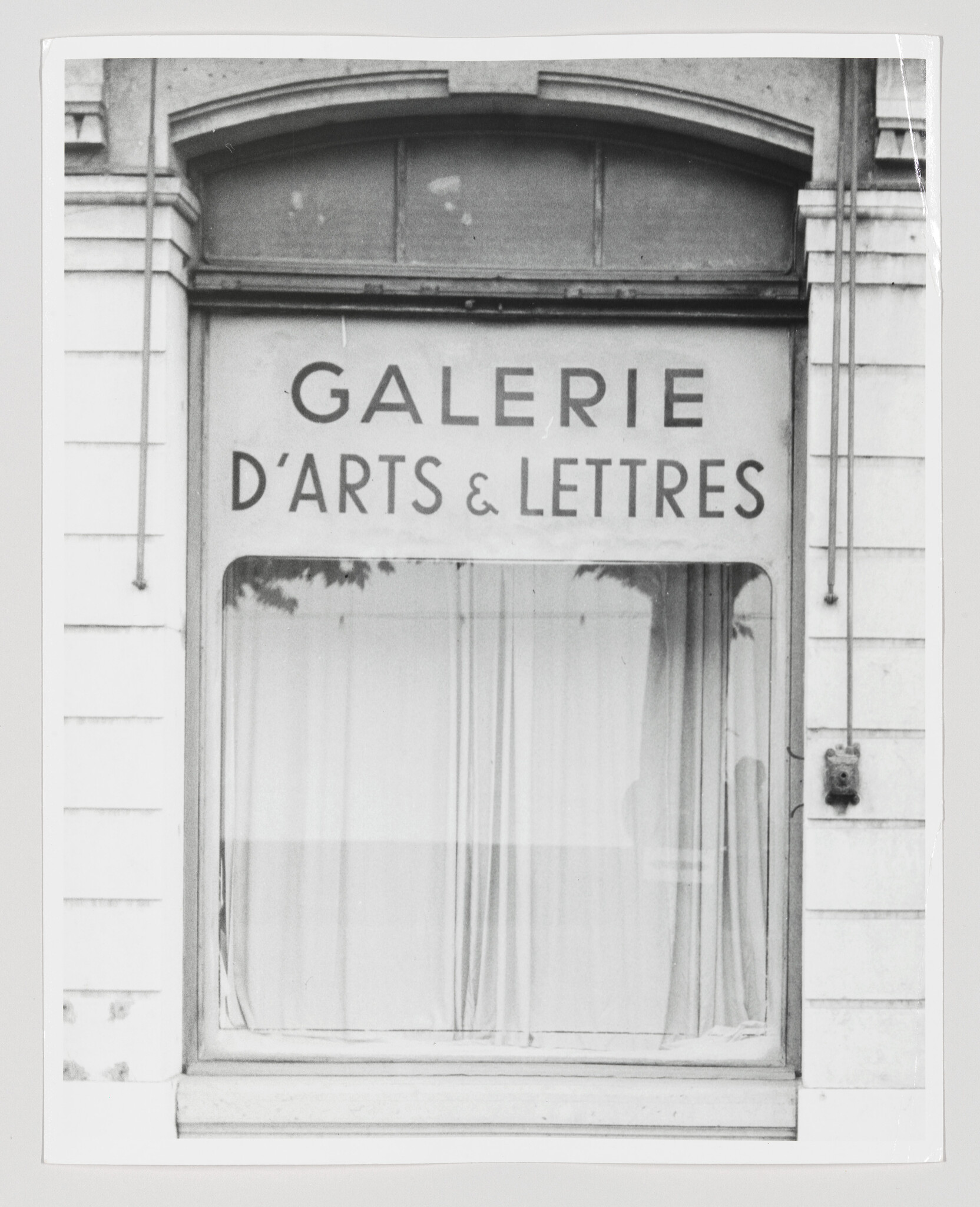 Windowed gallery storefront displays a sign reading "Galerie d'Arts & Lettres" above a curtained window.
