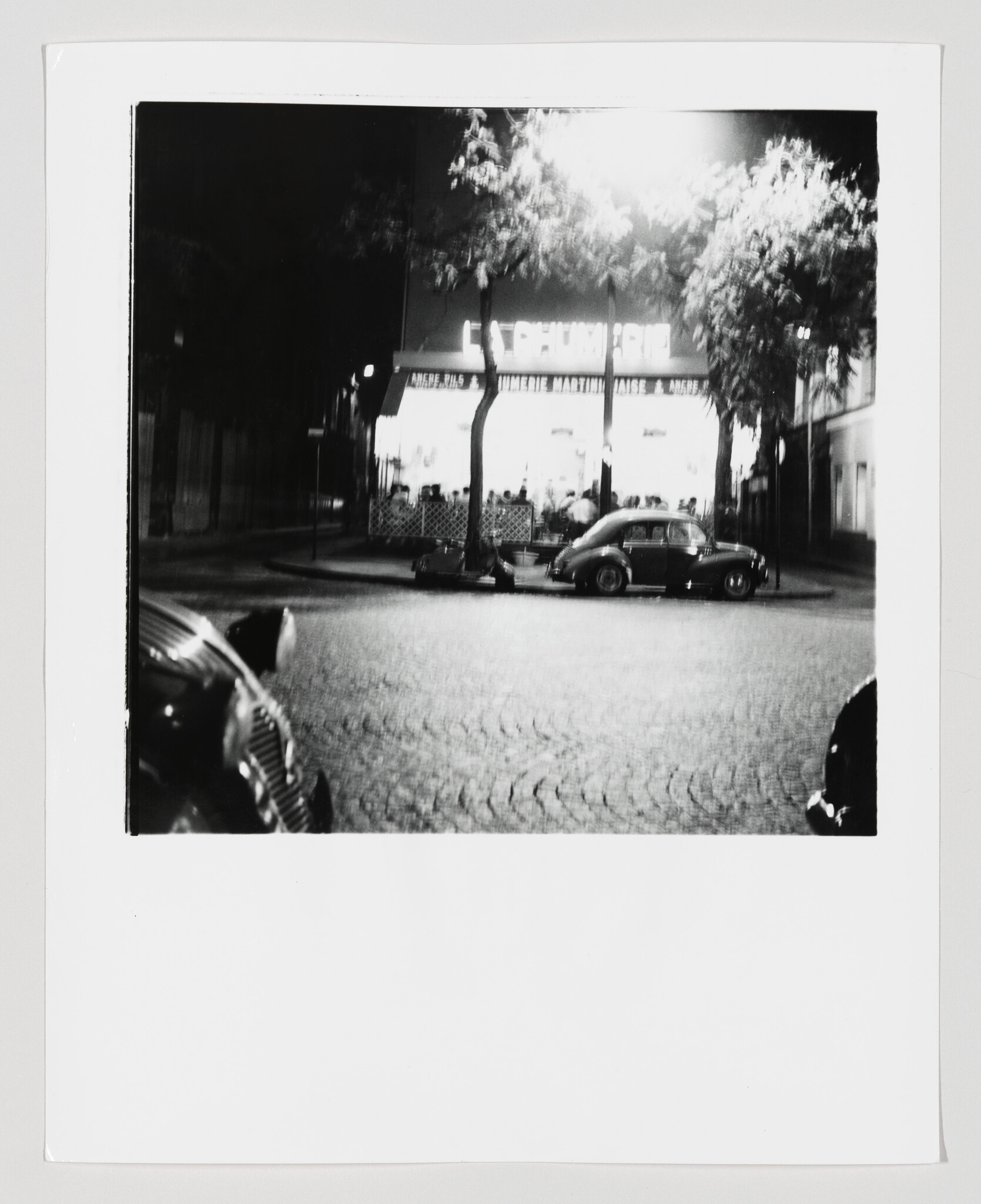 A lit sidewalk café at night with a vintage parked car and trees along the street.