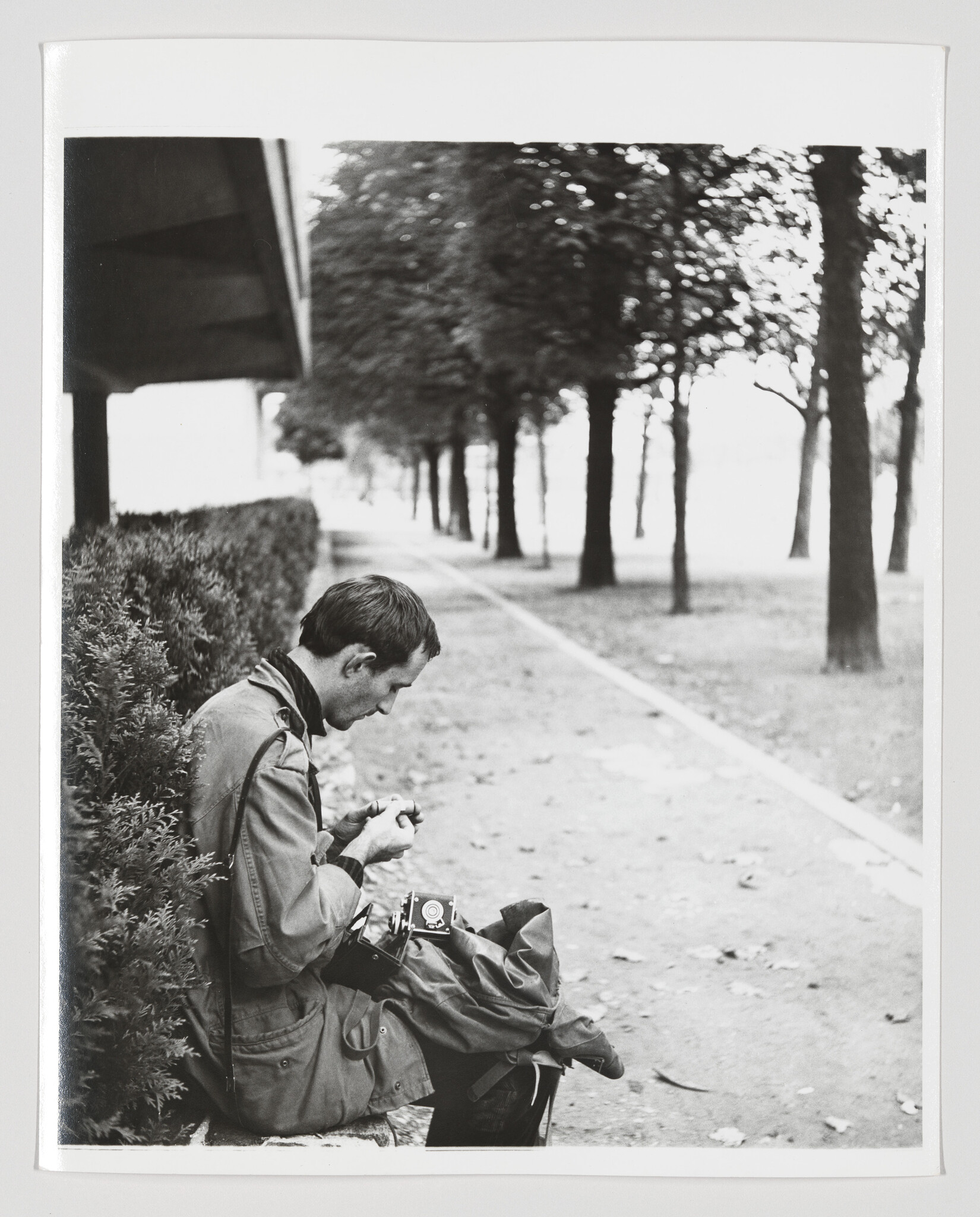 A man sits on a park bench adjusting his vintage camera while leaves scatter on the path.