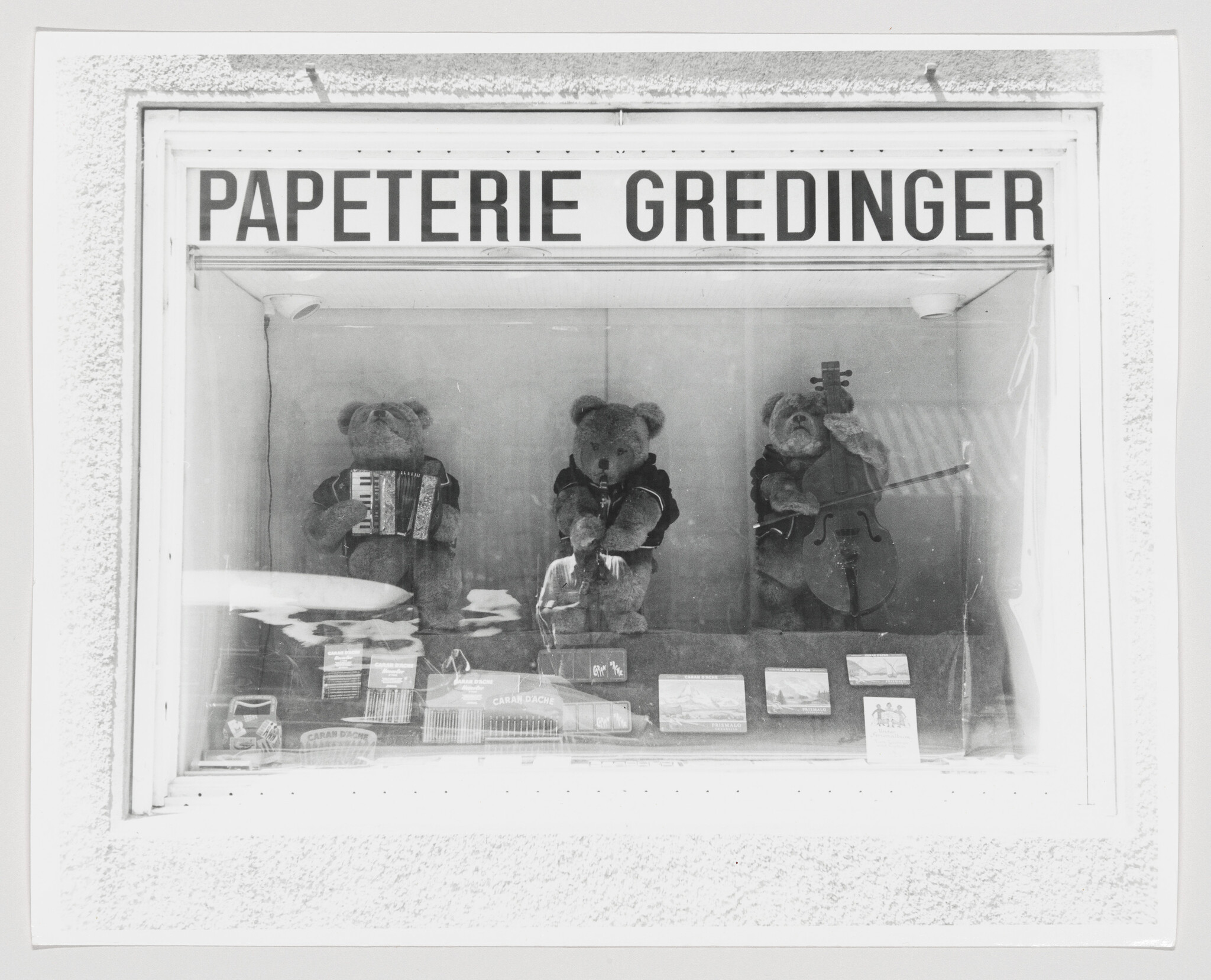 Three teddy bears wearing clothes play instruments in a store window labeled Papeterie Gredinger.