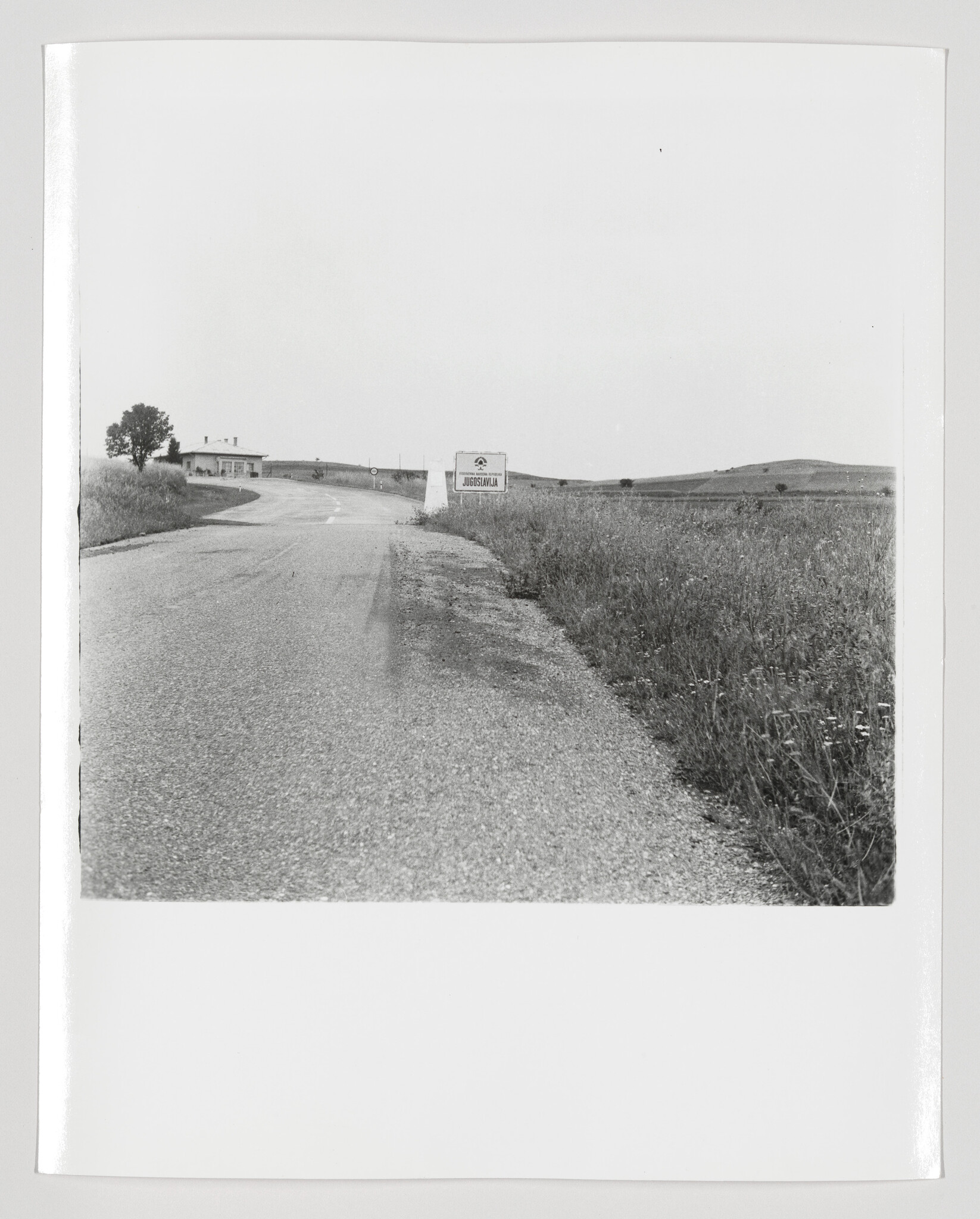 A country road curves toward a Yugoslavia border sign beside grassy fields.
