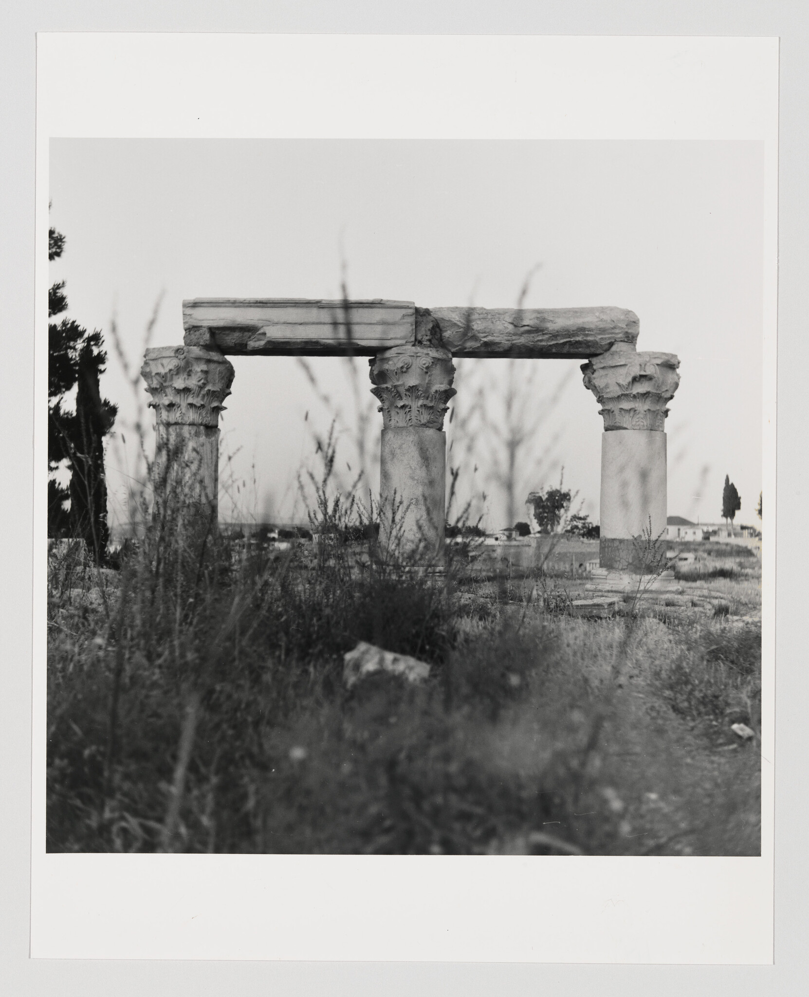 Three weathered ancient stone columns with carved capitals support a broken horizontal lintel amid overgrown grass.