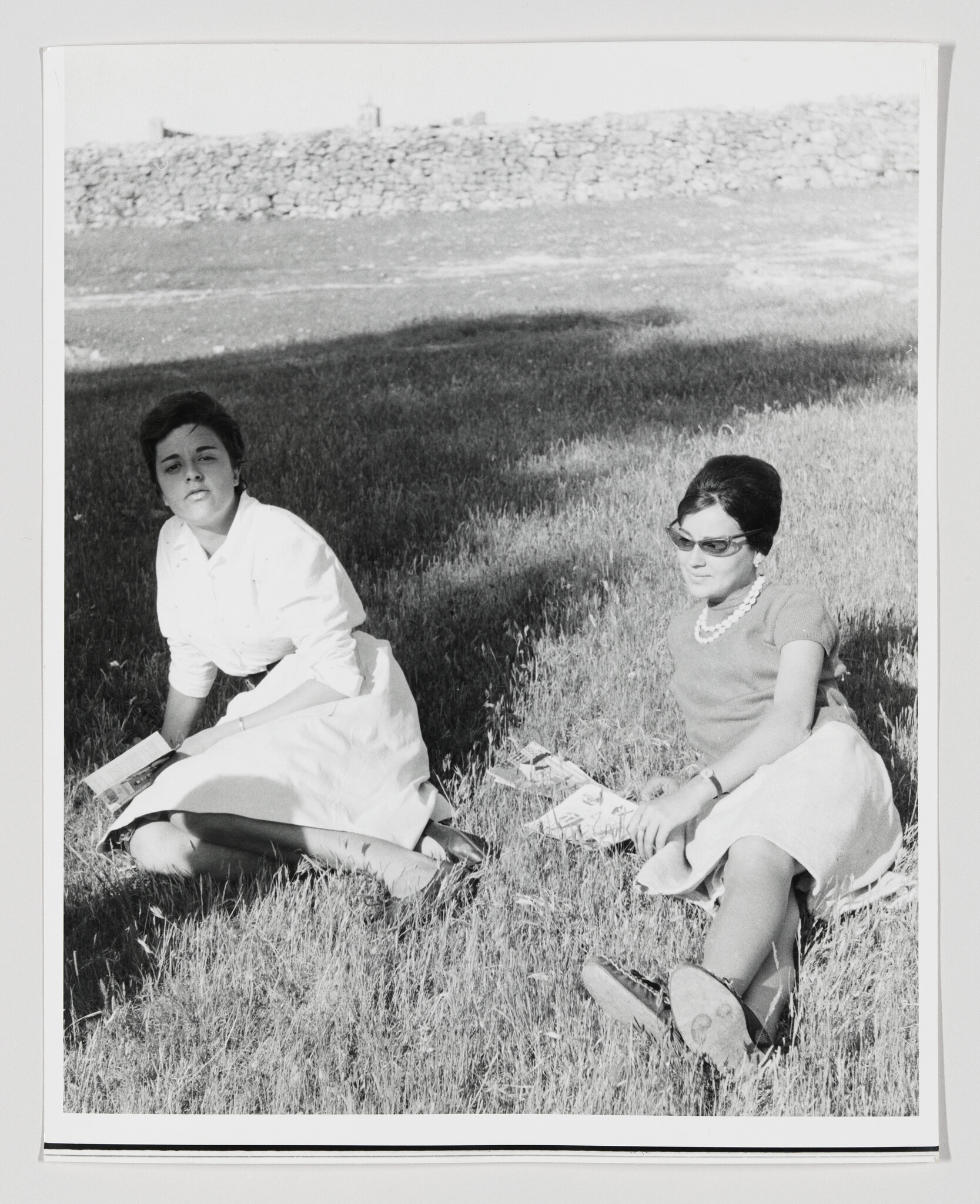 Two young women sit on the grass reading magazines and relaxing in a sunny field.