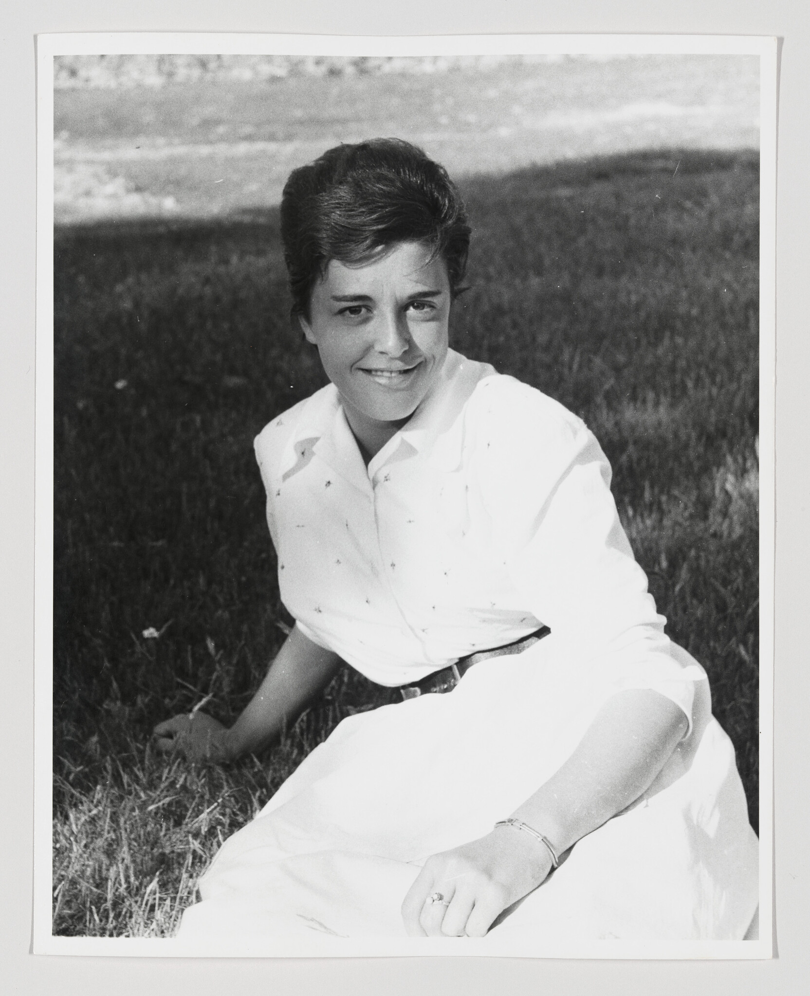 A young woman sits on grass smiling while wearing a white dress and a ring and bracelet.