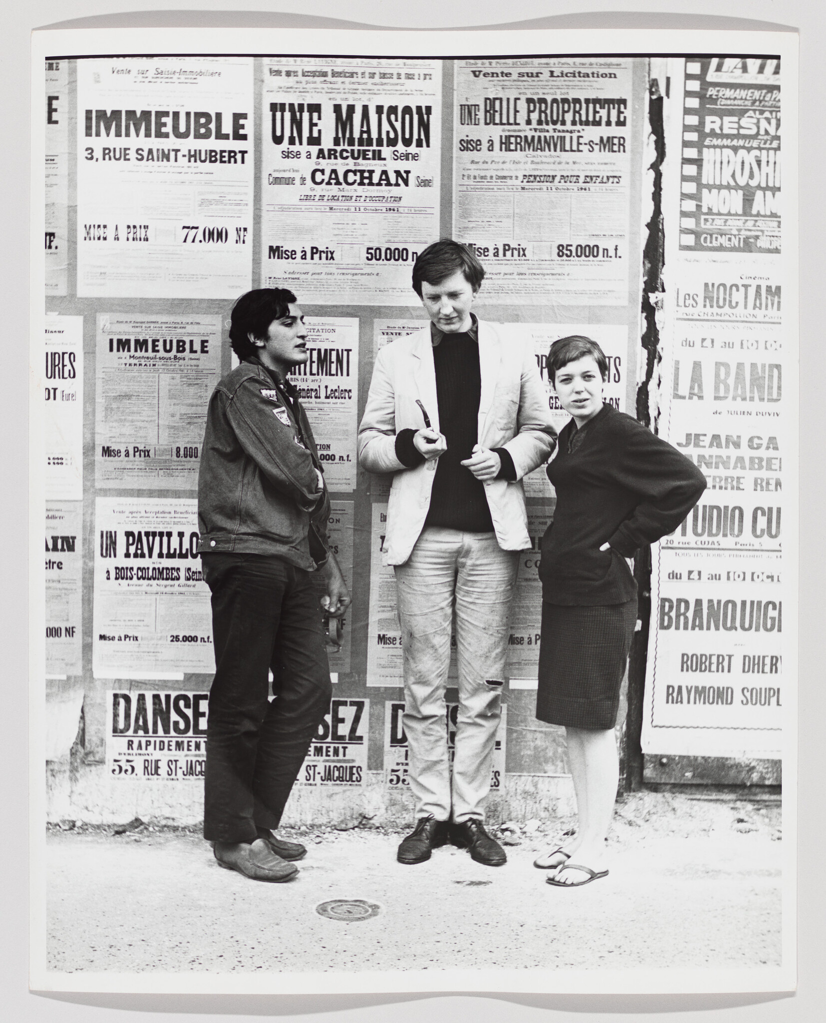 Three young people stand talking in front of a wall covered with French posters.