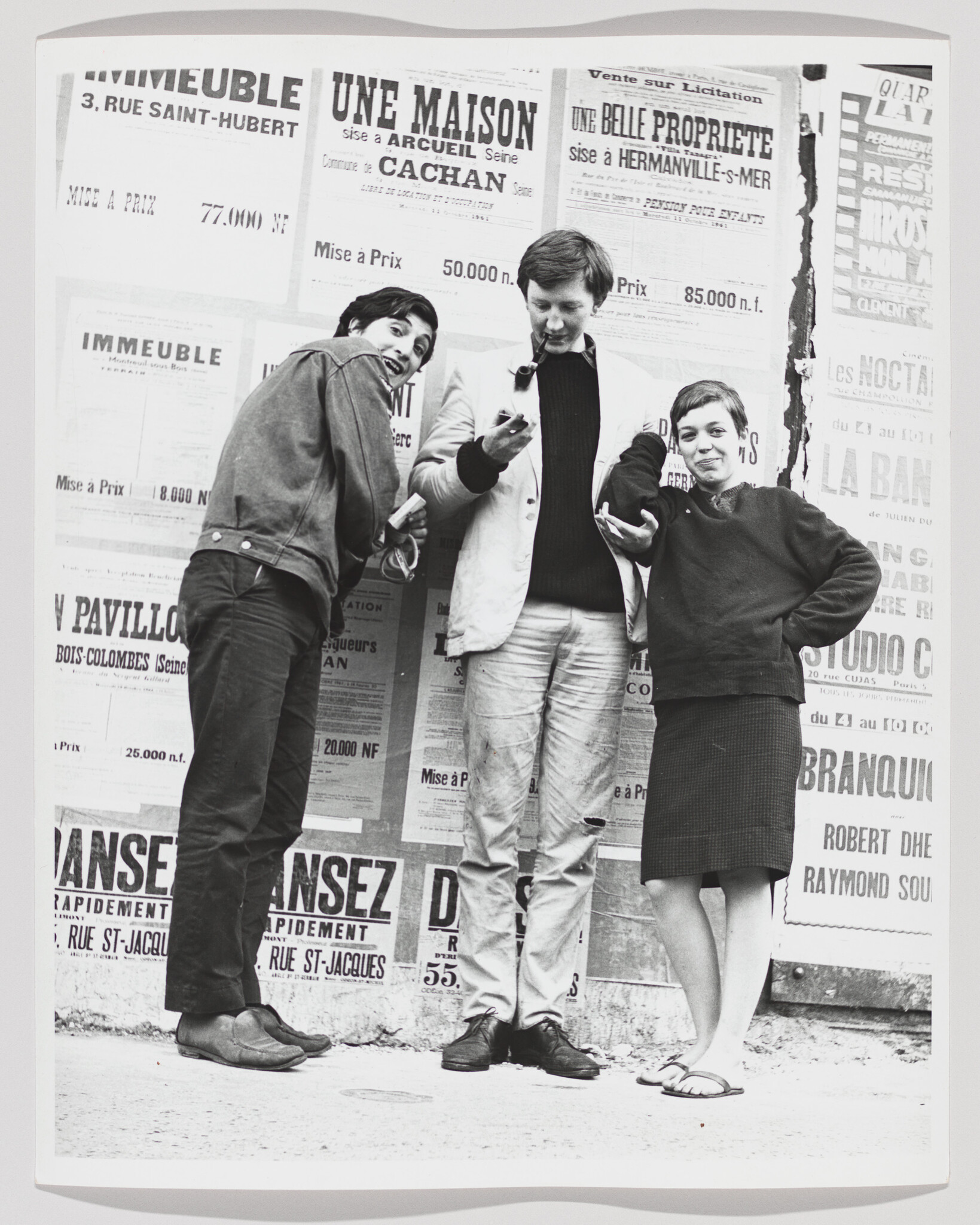 Three young people stand in front of a poster-covered wall, one smoking a pipe and others gesturing.