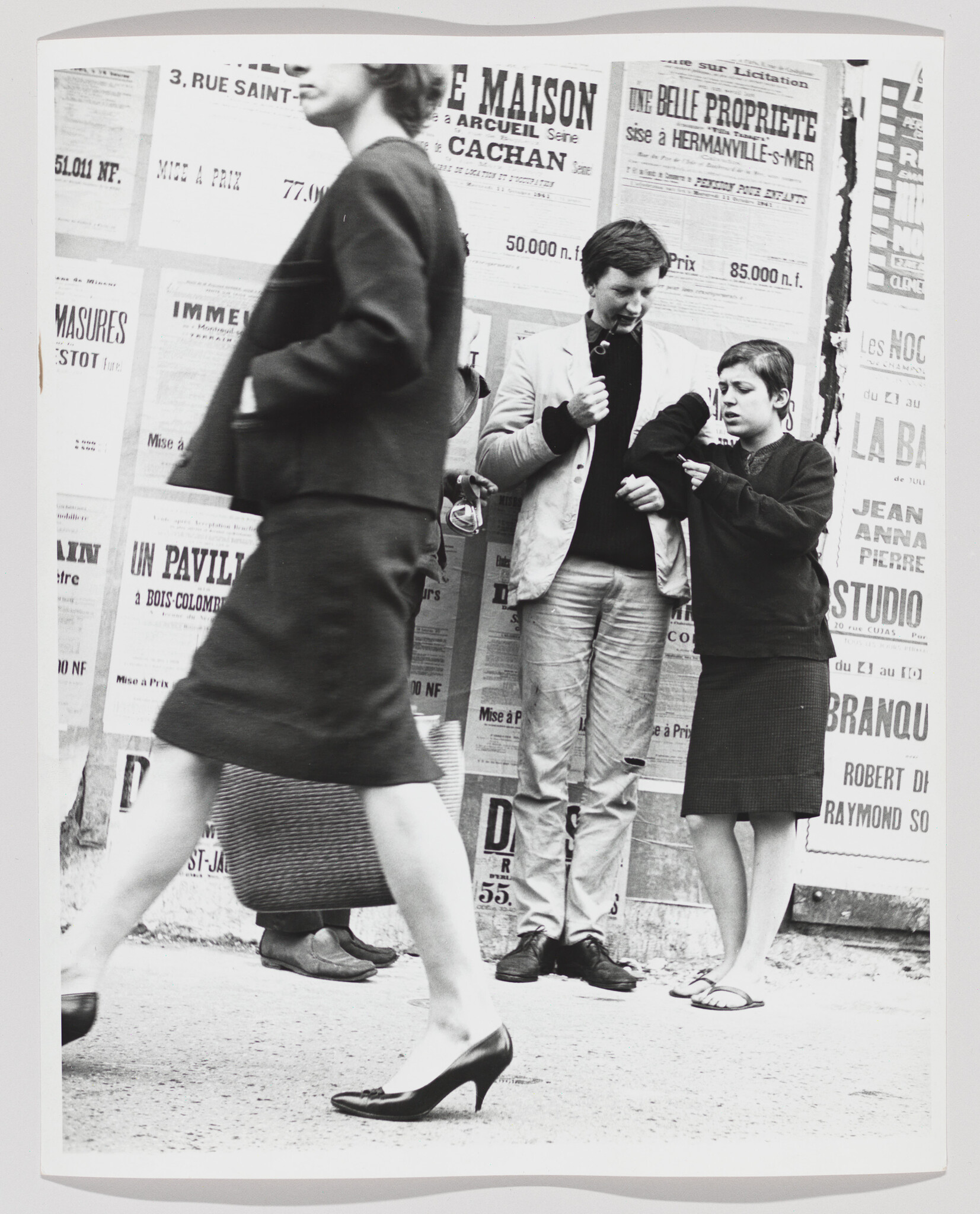 A woman strides past two teenagers examining something by a wall of posters.