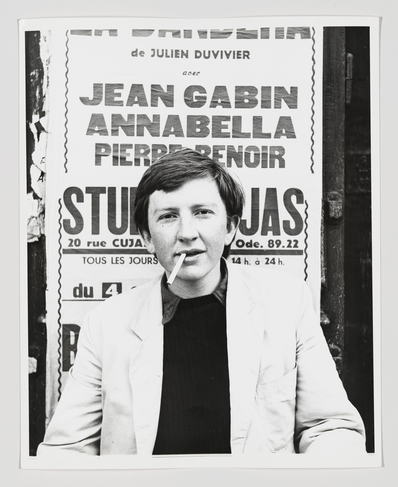 A young person in a light jacket holds a cigarette while sitting in front of a poster.