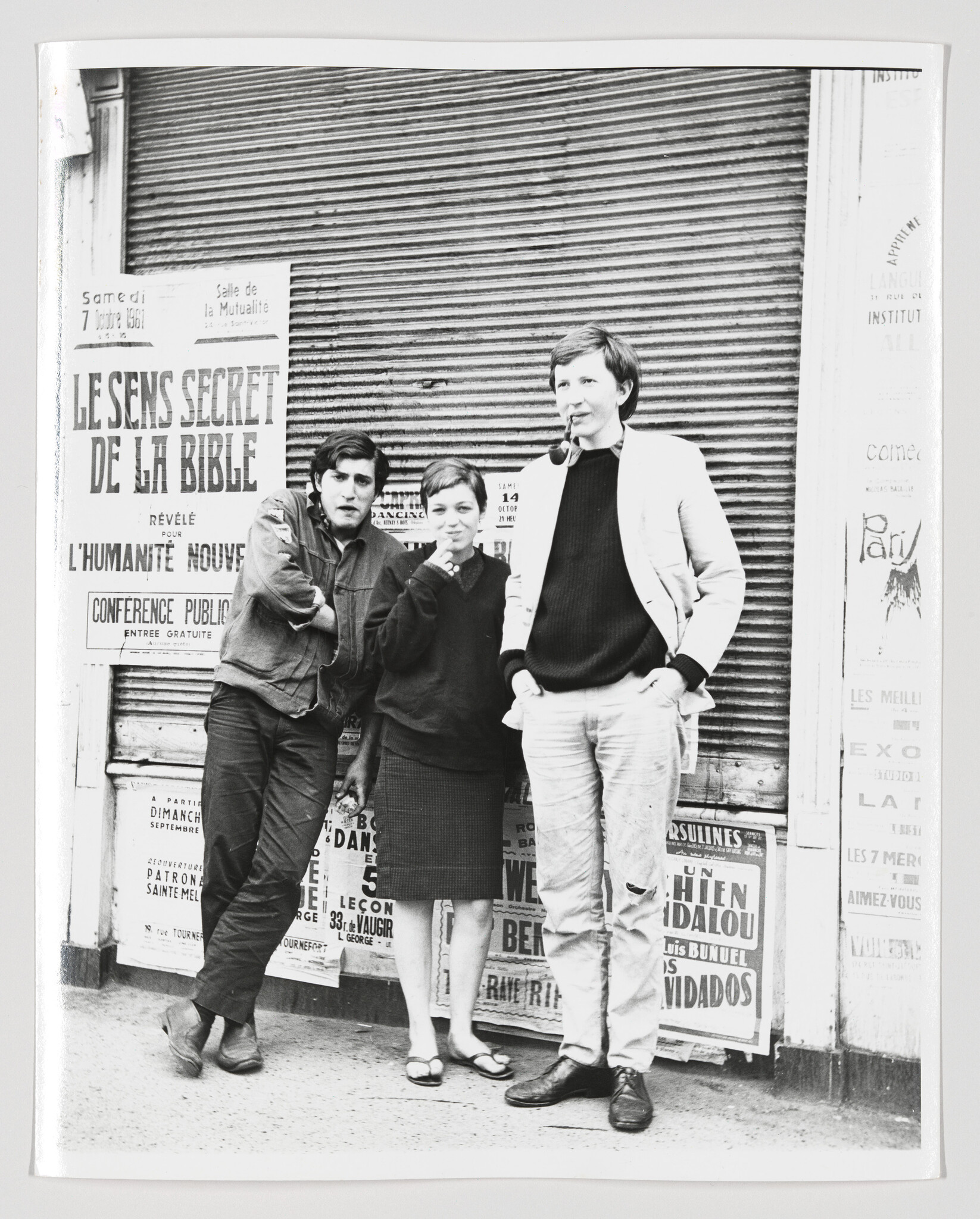 Three young adults stand in front of a closed shop shutter covered with various posters, including one that reads "LE SENS SECRET DE LA BIBLE." The two men and one woman appear casually dressed in styles indicative of mid-20th-century fashion.
