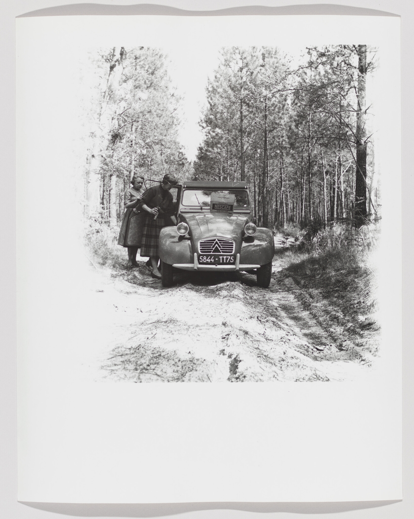 Two women examine a vintage Citroën car stopped on a sandy forest track.