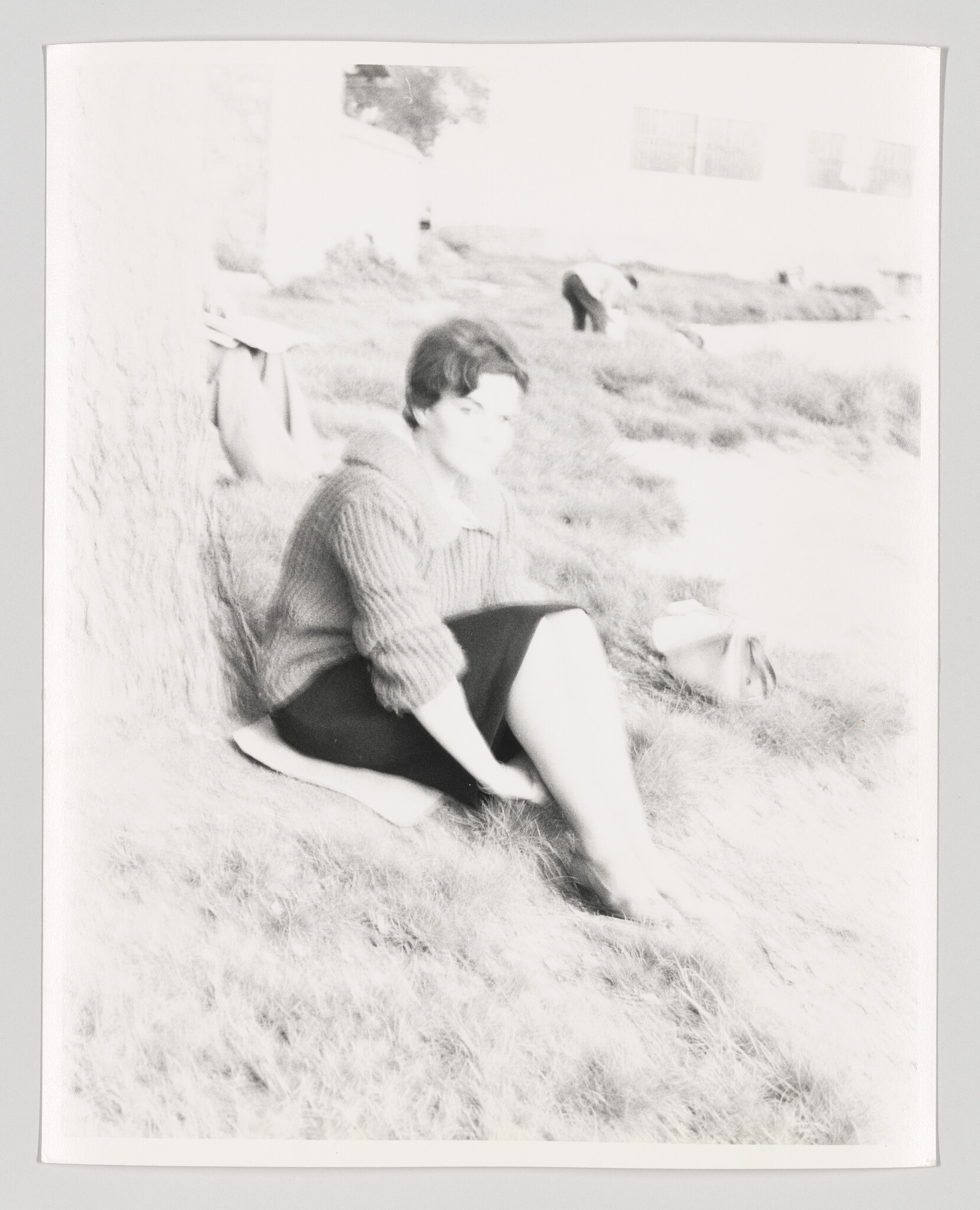 A woman in a sweater sits on the grass under a tree with a bag nearby.
