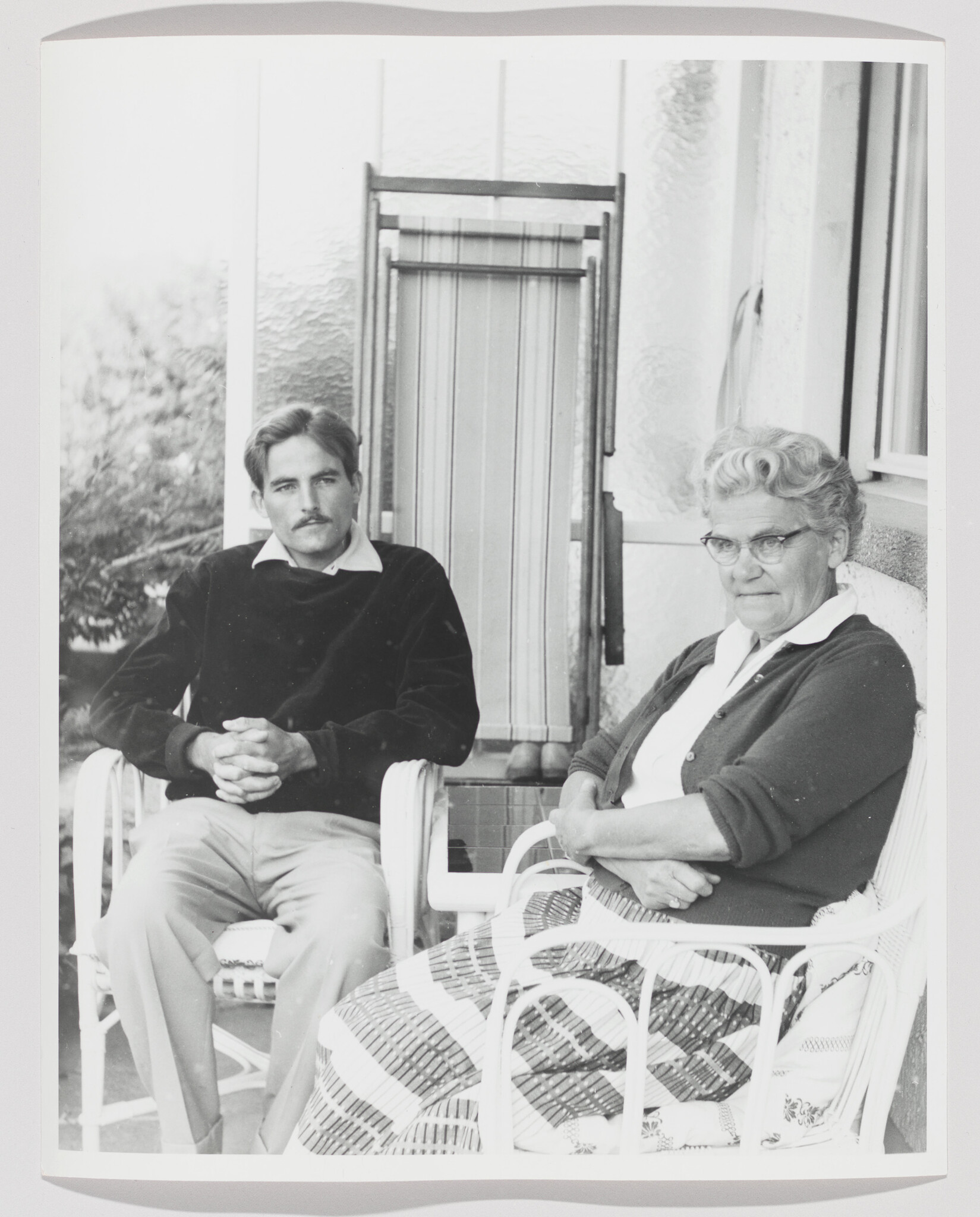 An older woman and a younger man sit on wicker chairs on a porch, looking at the camera.