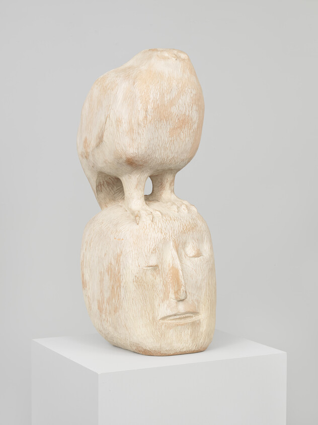 Carved pale wooden sculpture of a human head with a bird perched on top, legs touching the forehead.