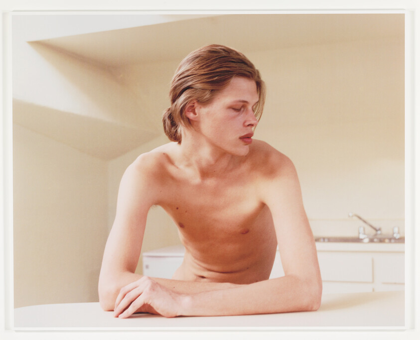 A shirtless young person leans on a kitchen counter, looking to the right.