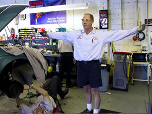 A mechanic stands with arms outstretched while another person works under a raised car in a garage.