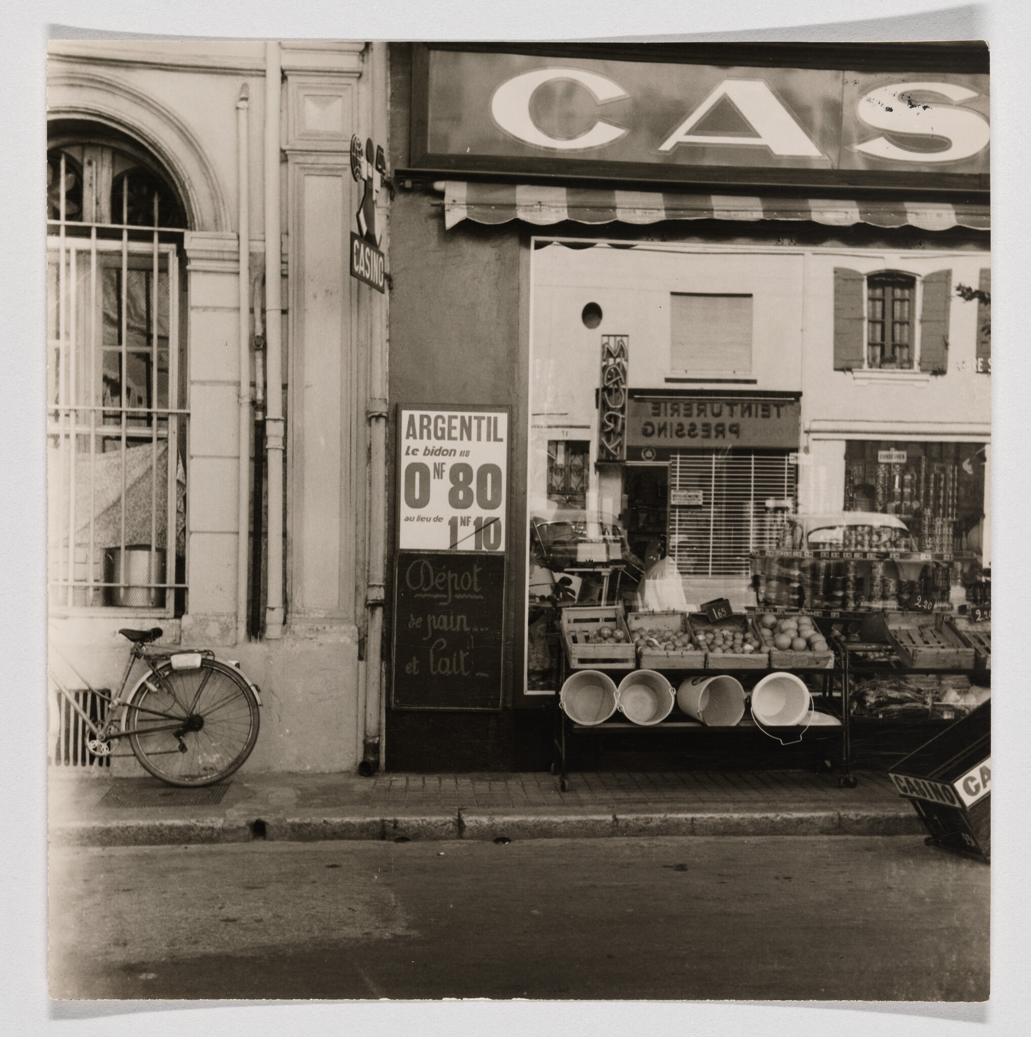 Grocery storefront with fruit crates and white buckets displayed on the sidewalk under a striped awning.