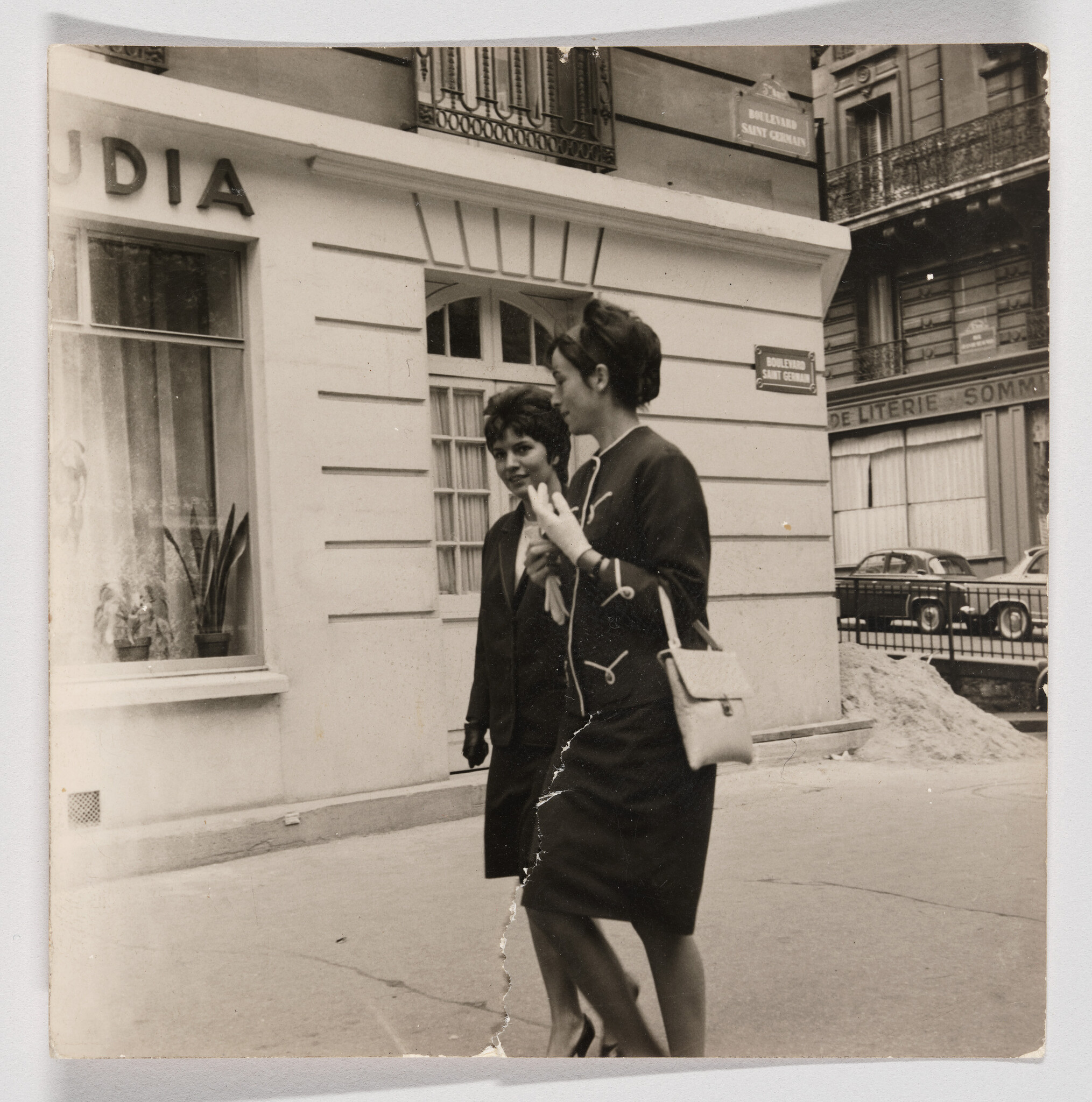 Two women walk together on a city sidewalk, one carrying a small handbag and talking.