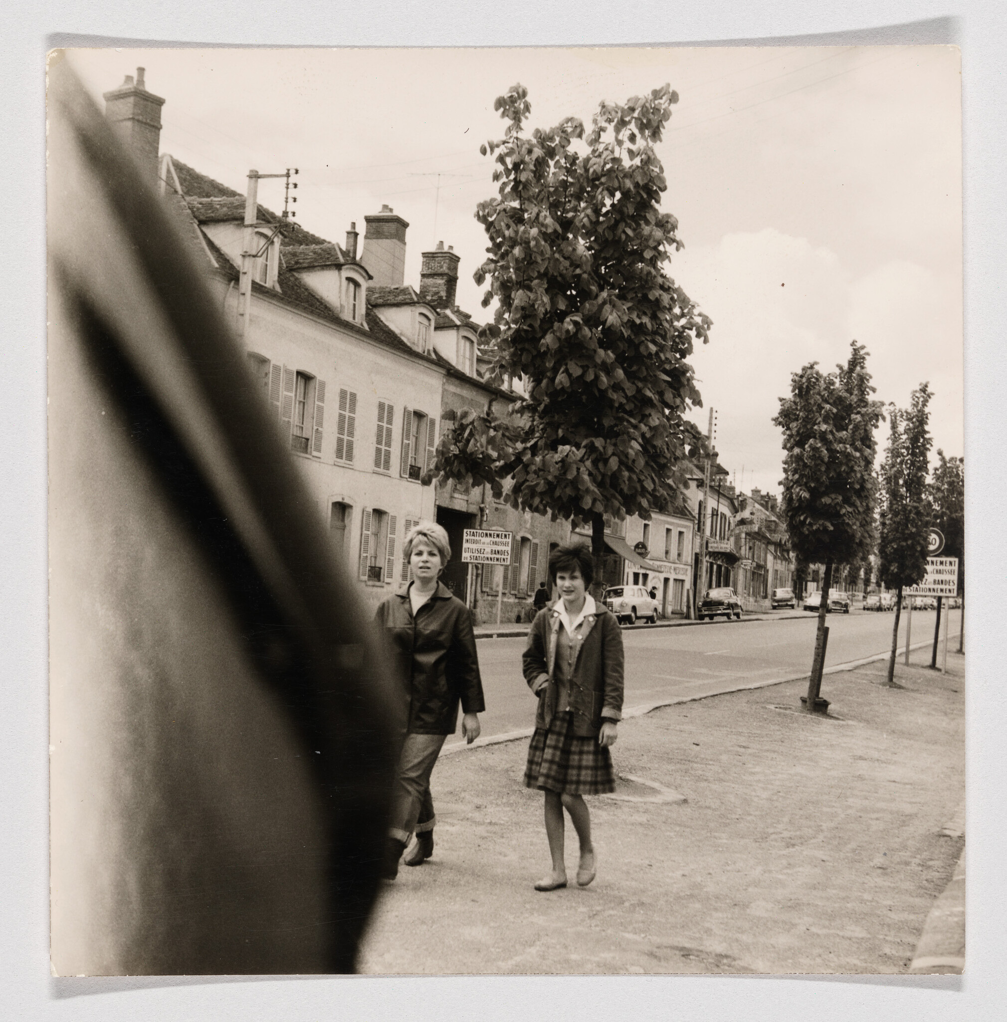 Two young women walking along a quiet street lined with trees and old buildings.