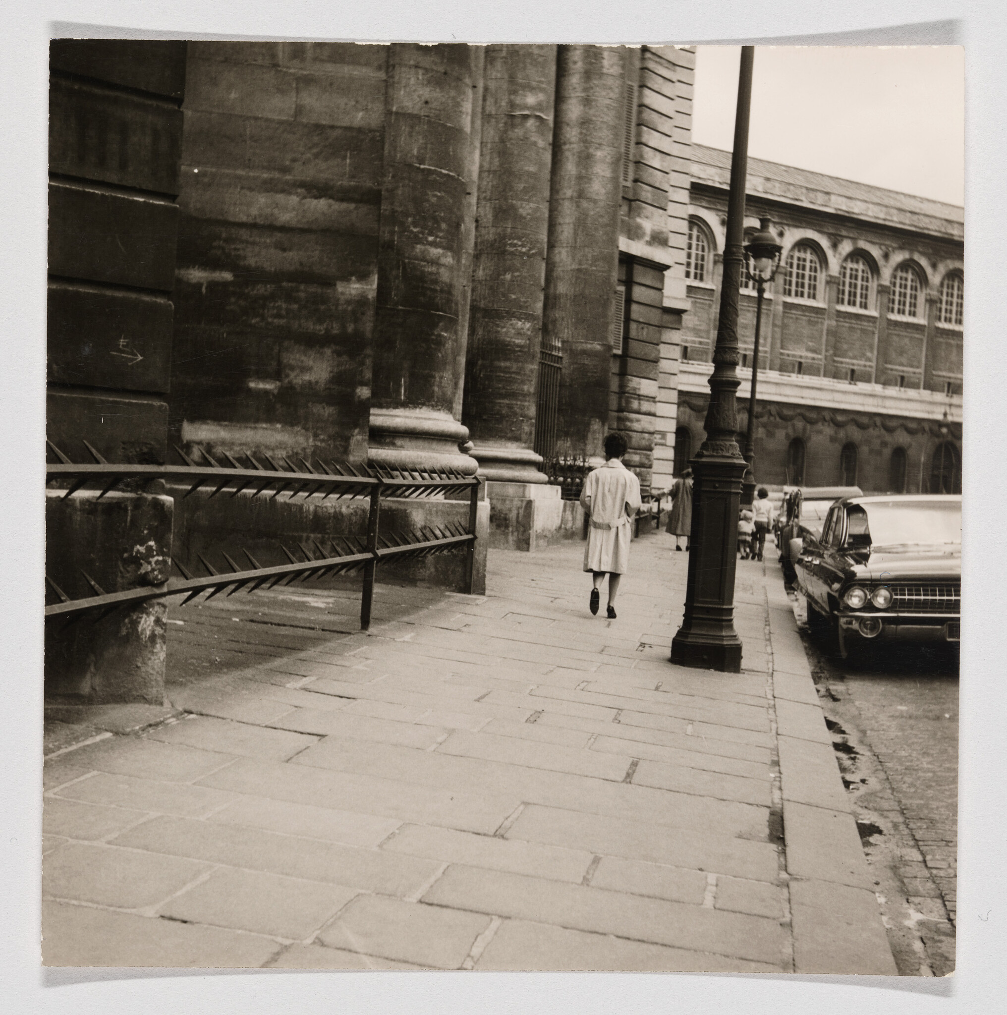 A person in a light coat walks away along a wide sidewalk beside a stone building.